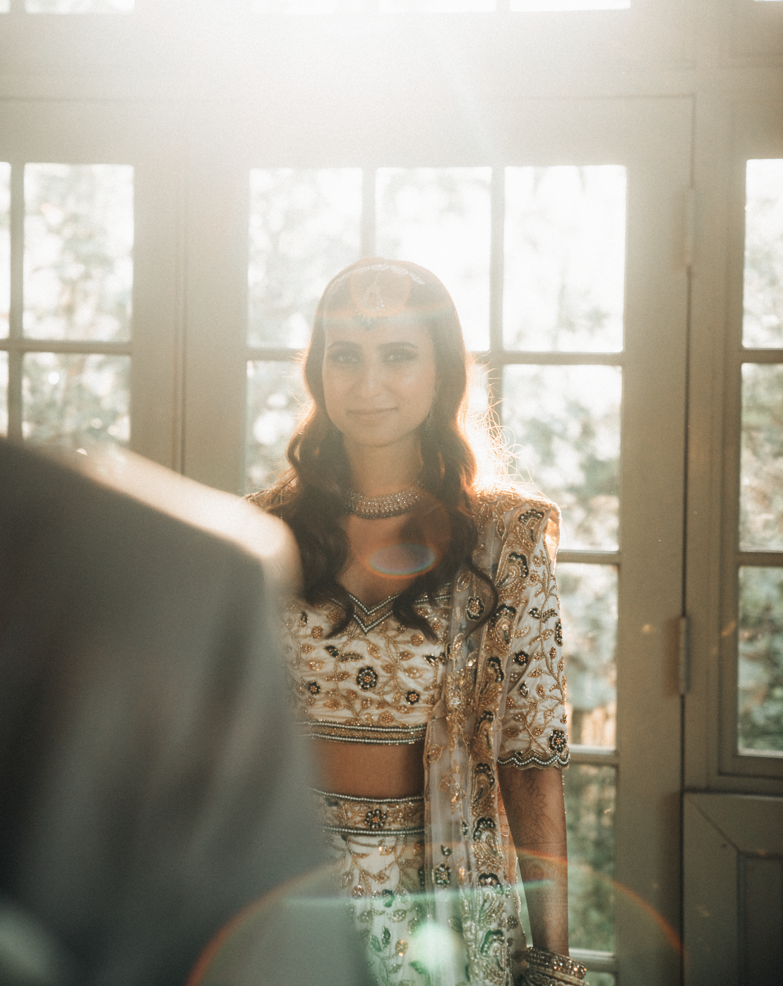 A woman dressed in traditional Indian attire standing near a sunlit window with trees outside.