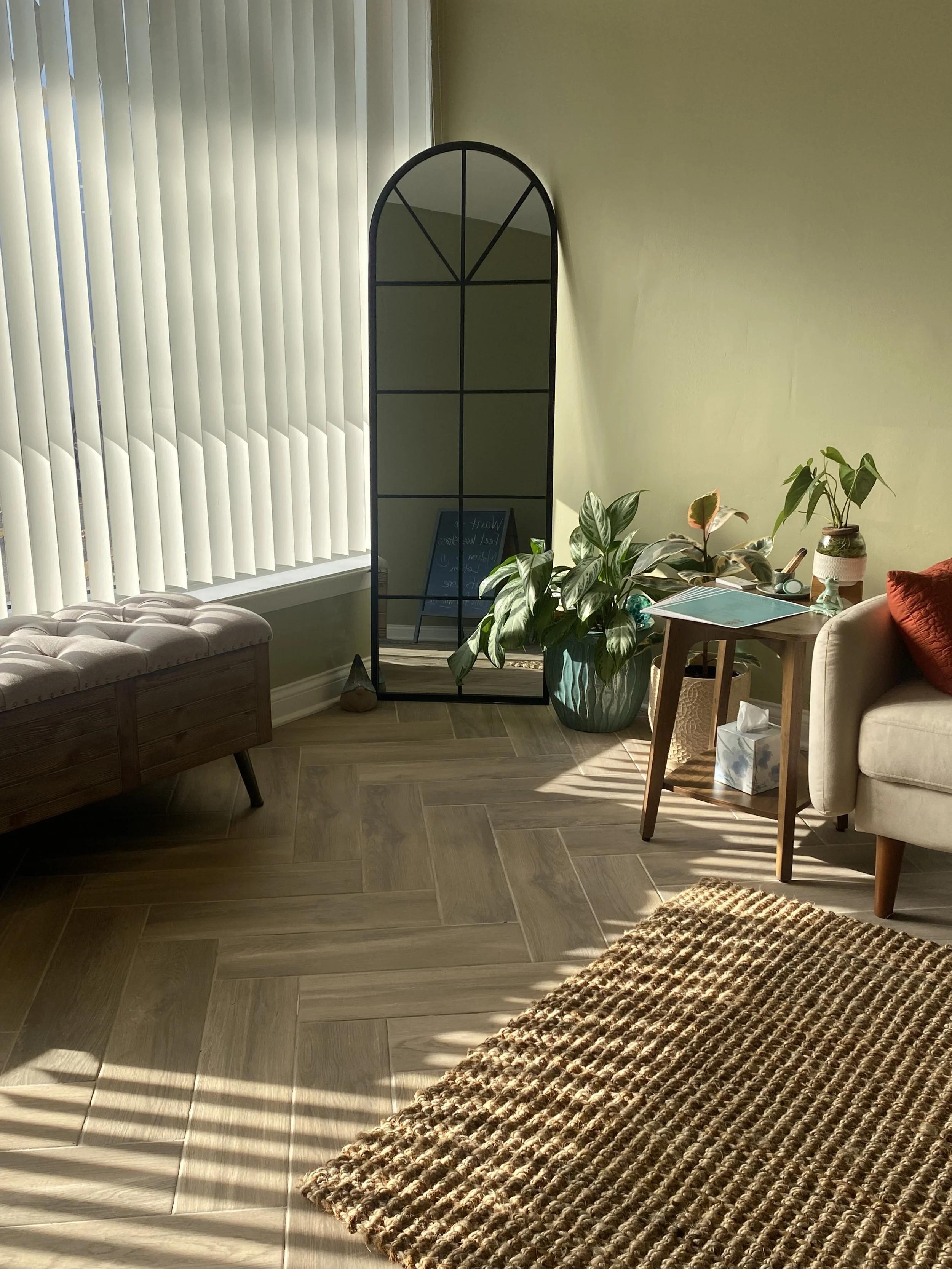Sunlit living room corner with wooden floor, a beige tufted bench, a large potted green plant, a small wooden side table with a tissue box, a white armchair with an orange pillow, a tall black metal mirror, and vertical blinds on a window casting shadows on the floor.