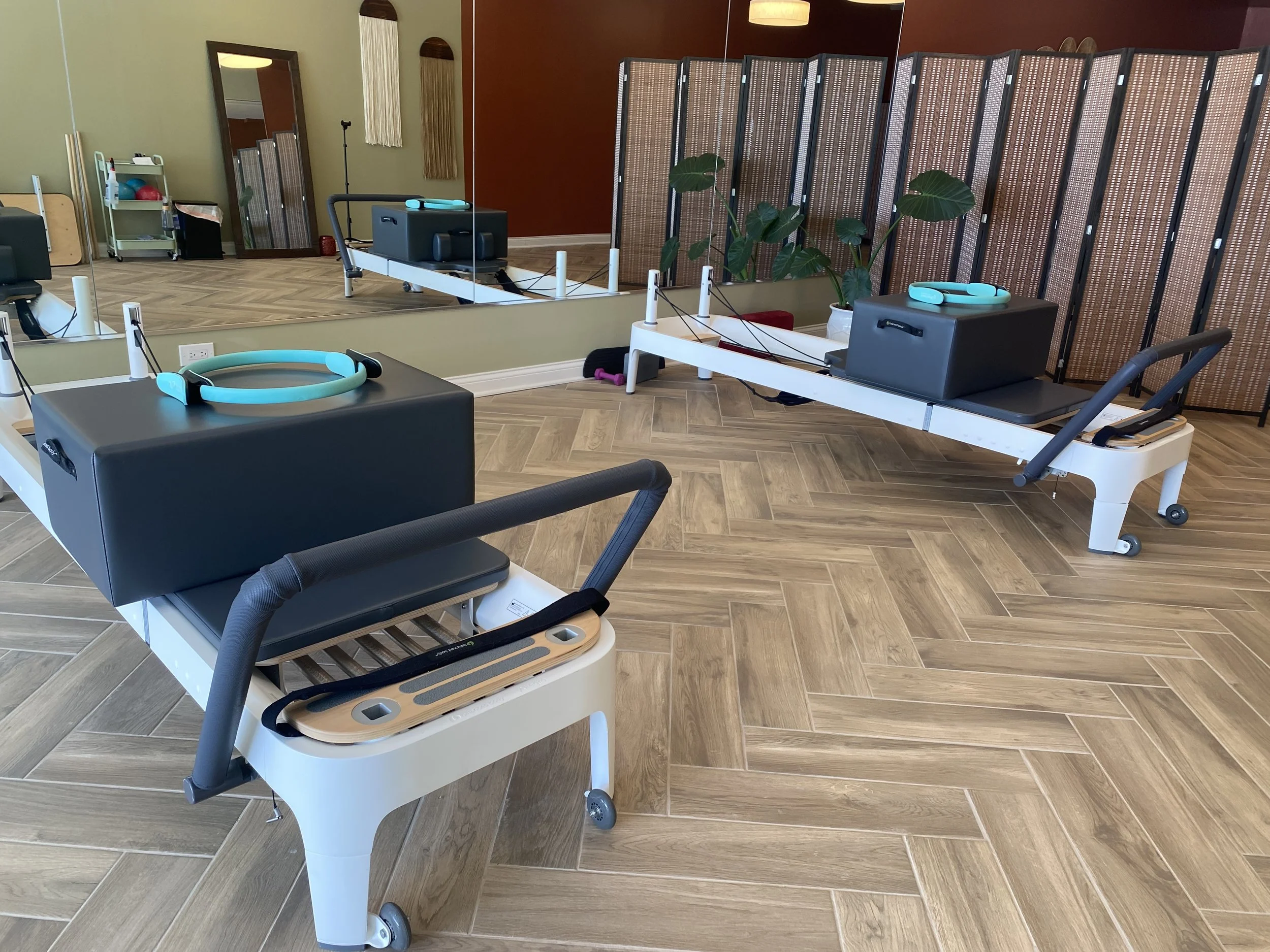 Two Pilates reformer machines in a studio with large mirror and plants, set on wood-look tile floor, with a room divider and various fitness equipment in the background.