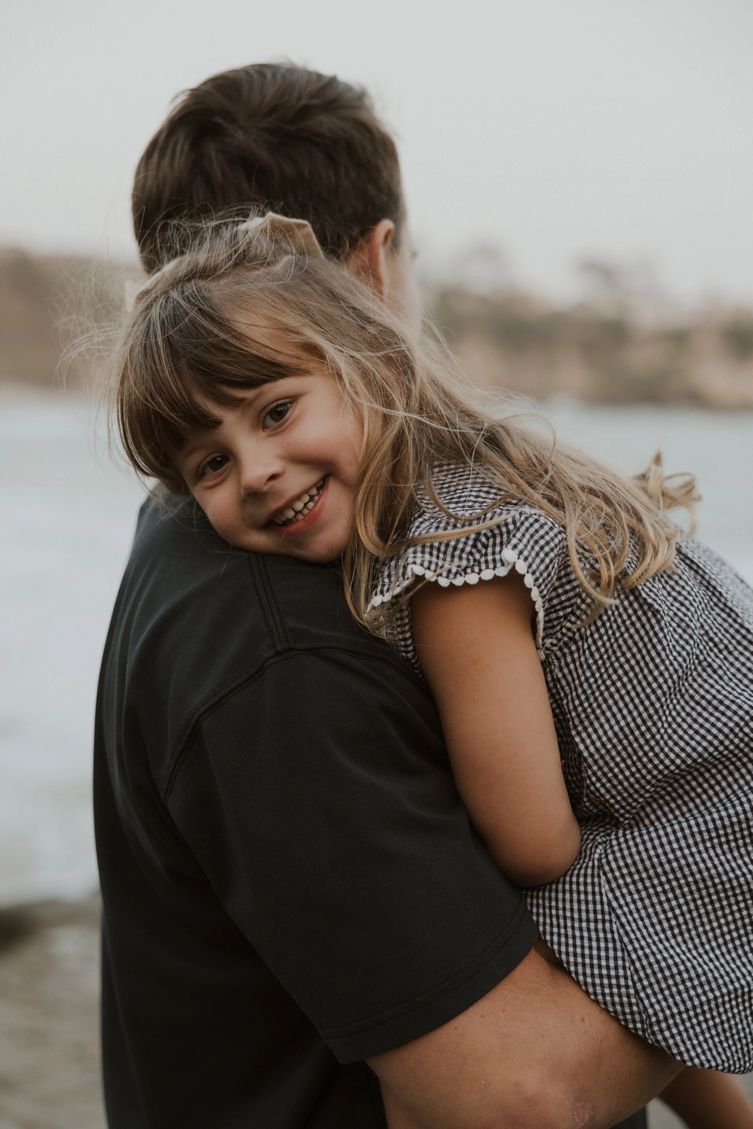 A young girl smiling while being carried on someone's shoulder by the seaside.