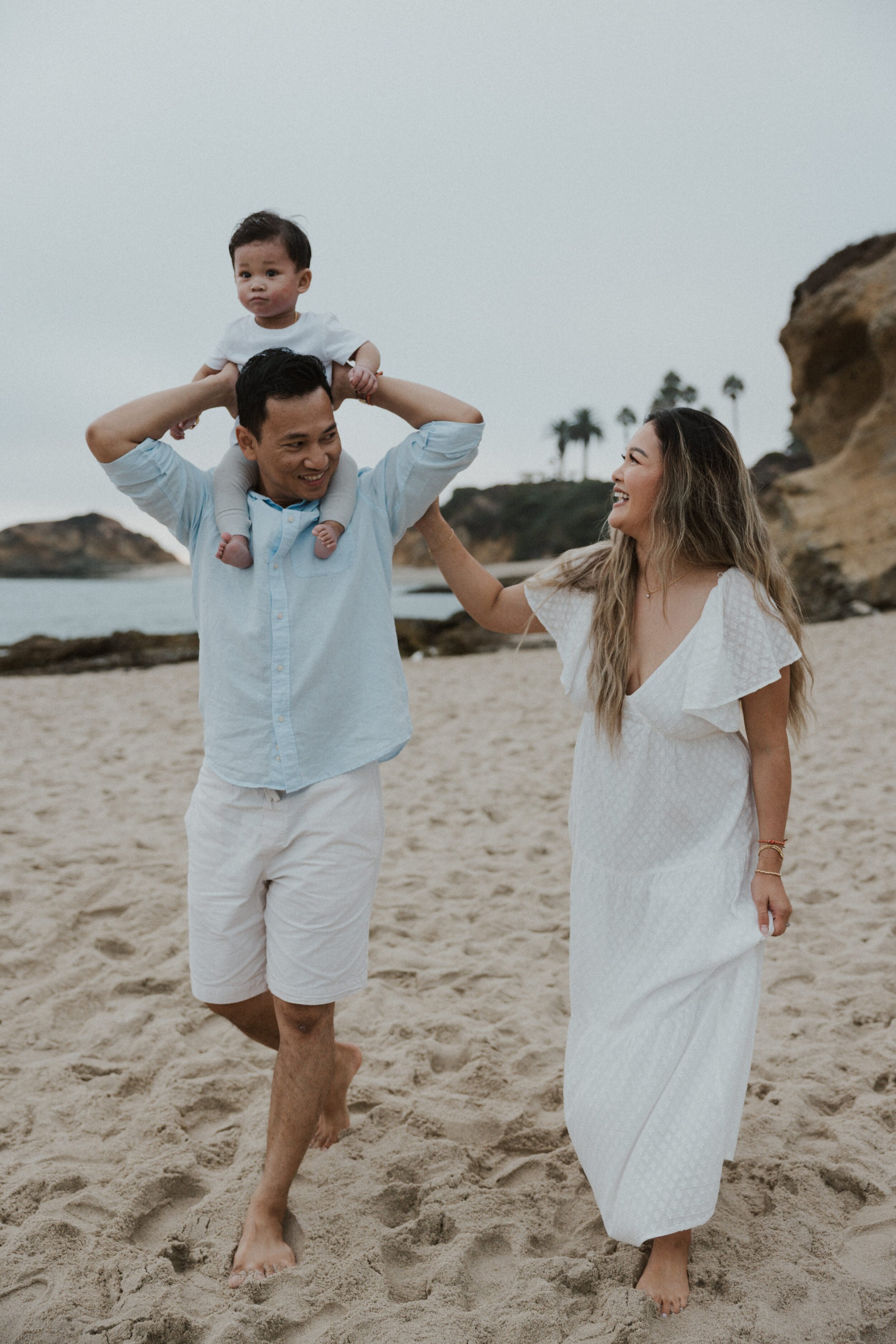 A family walking on a beach, with a man carrying a baby on his shoulders and a woman smiling at them.
