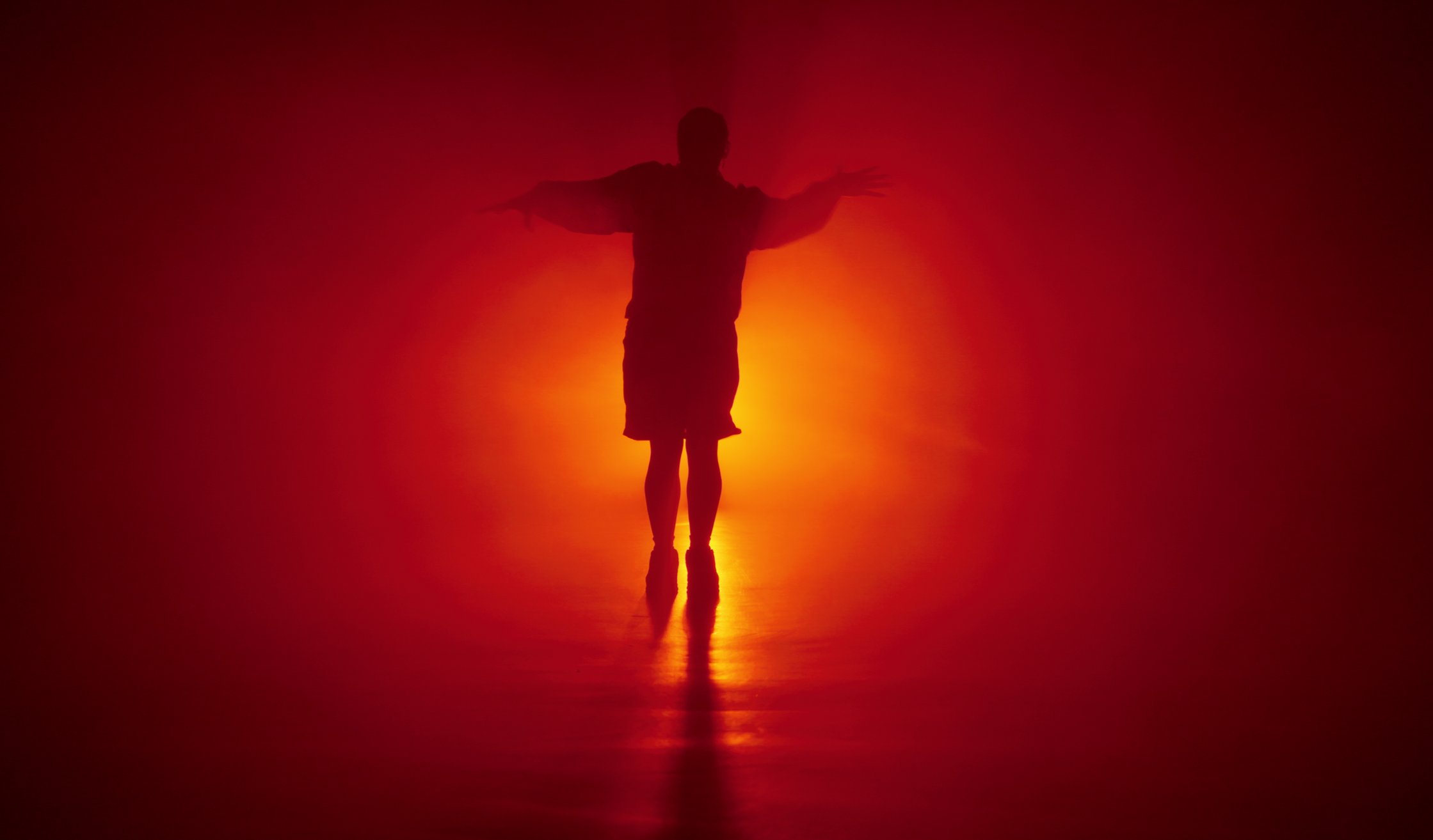 Silhouette of a person standing with arms outstretched against a red and orange gradient background, reflections on the ground.