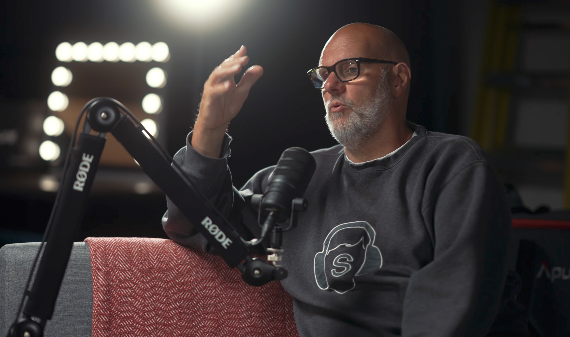 A man with a gray beard and glasses speaking into a microphone during a recording or podcast session, with studio lighting in the background.