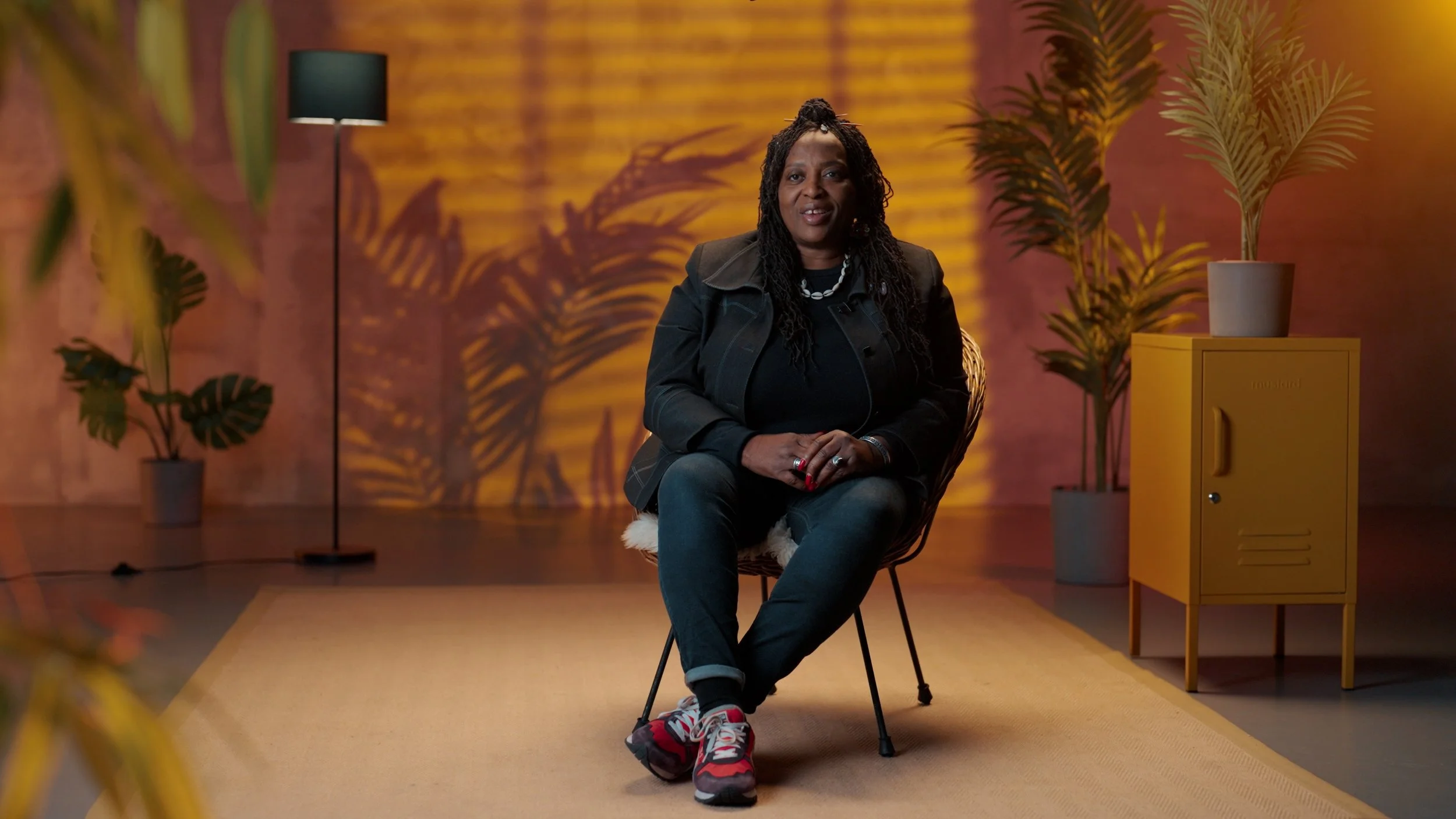 A woman with long dreadlocks sitting on a chair in a room with warm lighting, surrounded by houseplants and a yellow cabinet.