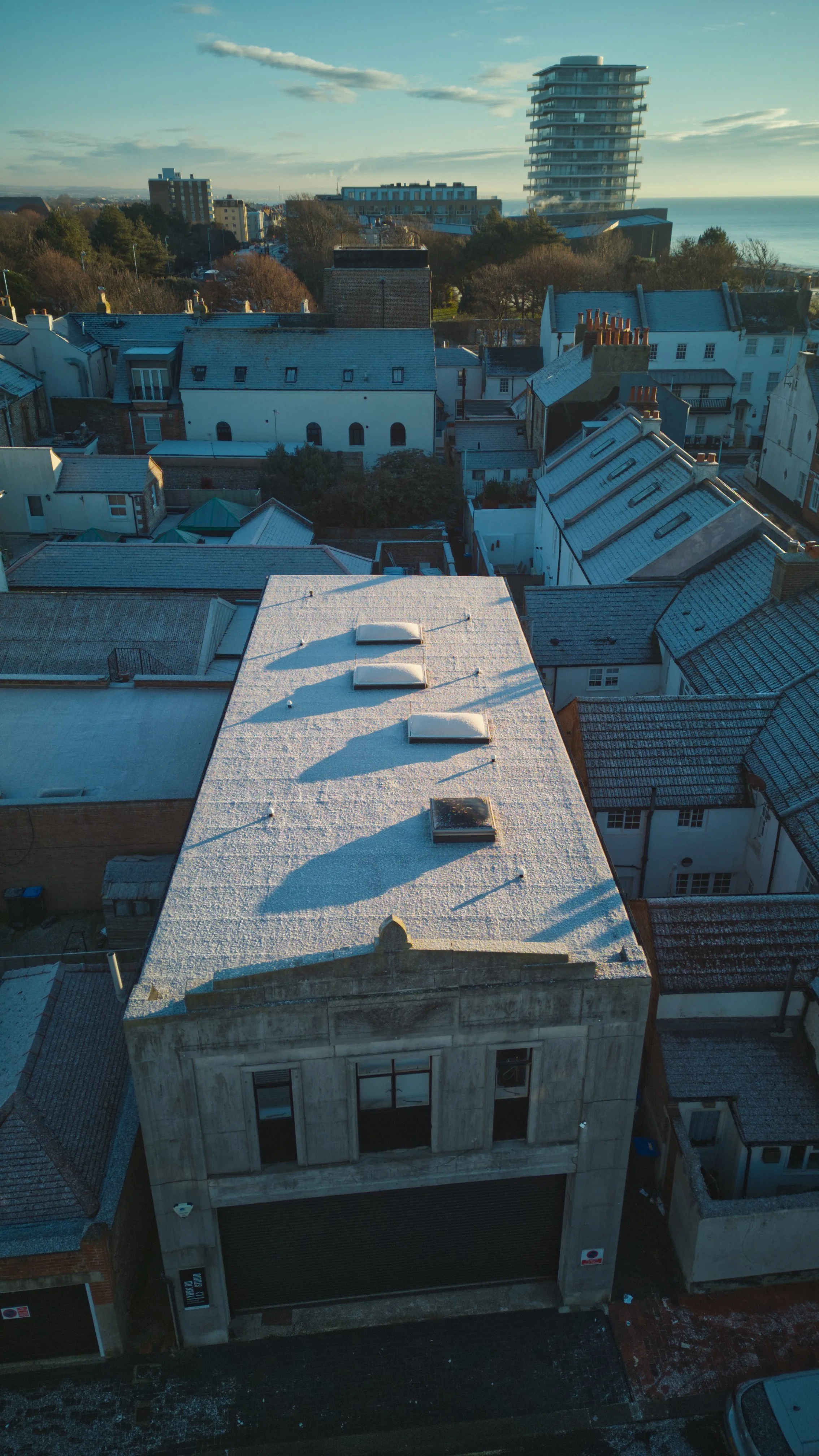 Aerial view of a city street with white and beige buildings, some with snow on their roofs, and a large modern building in the background near the ocean.