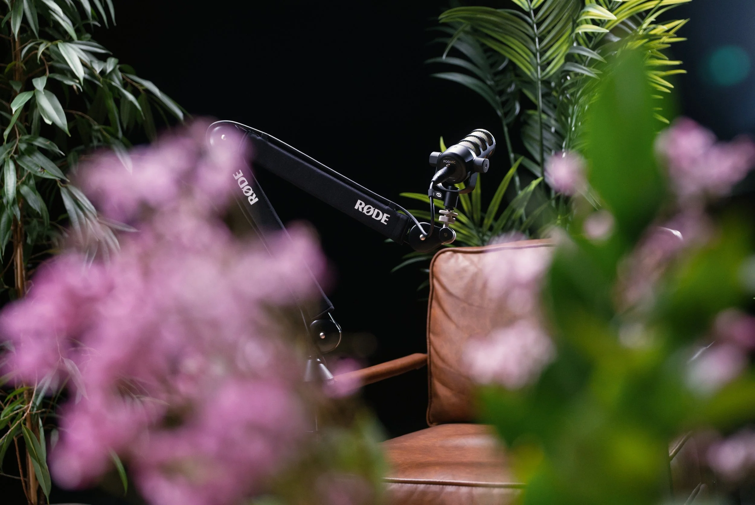 A microphone labeled 'Podmic' attached to a boom arm, surrounded by lush green foliage and pink flowers.