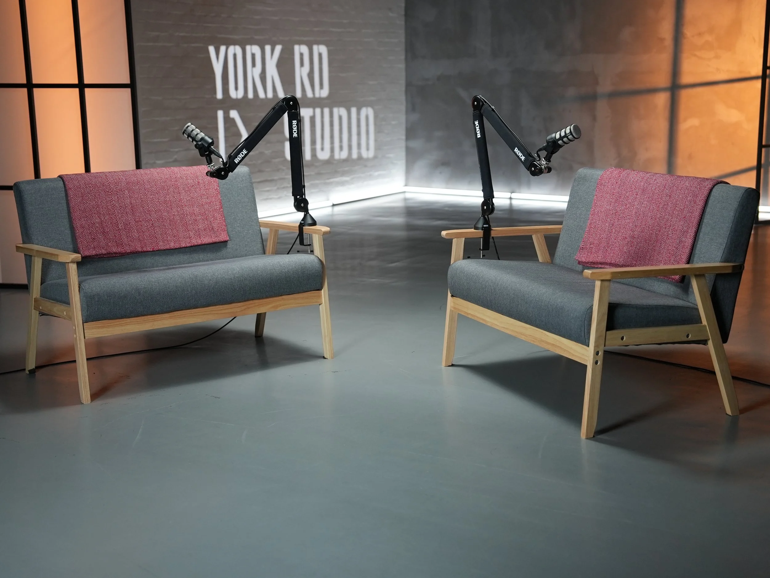 A podcast recording setup with two grey chairs with red back cushions, each equipped with a black Rode adjustable arm microphone. The background features a grey wall with white text reading 'YORK RD STUDIOS' and orange lighting on the sides.