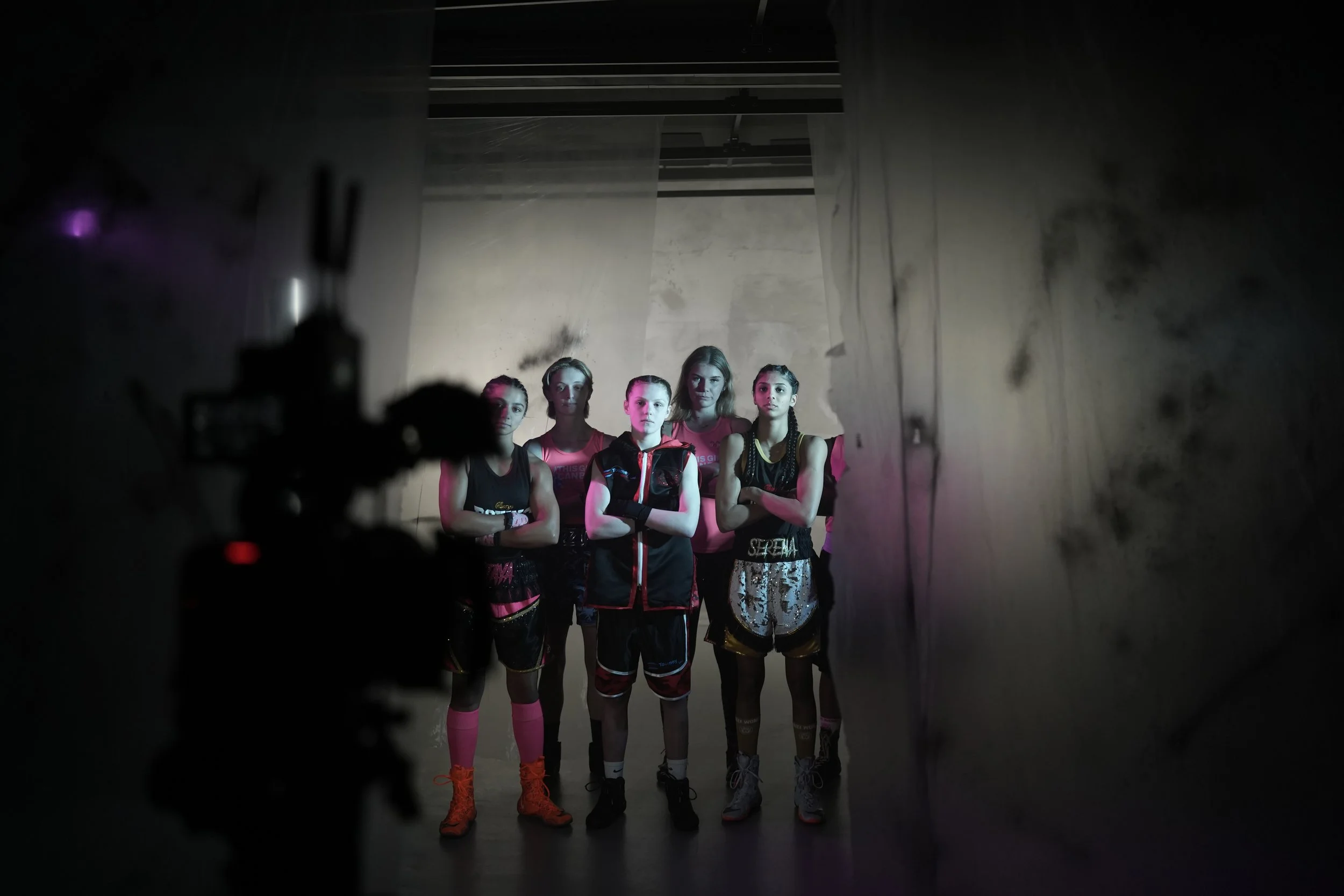 A group of five young women in athletic gear standing in a line in front of a plain textured wall, with the photo taken through an opening or behind a curtain, creating a framing effect.