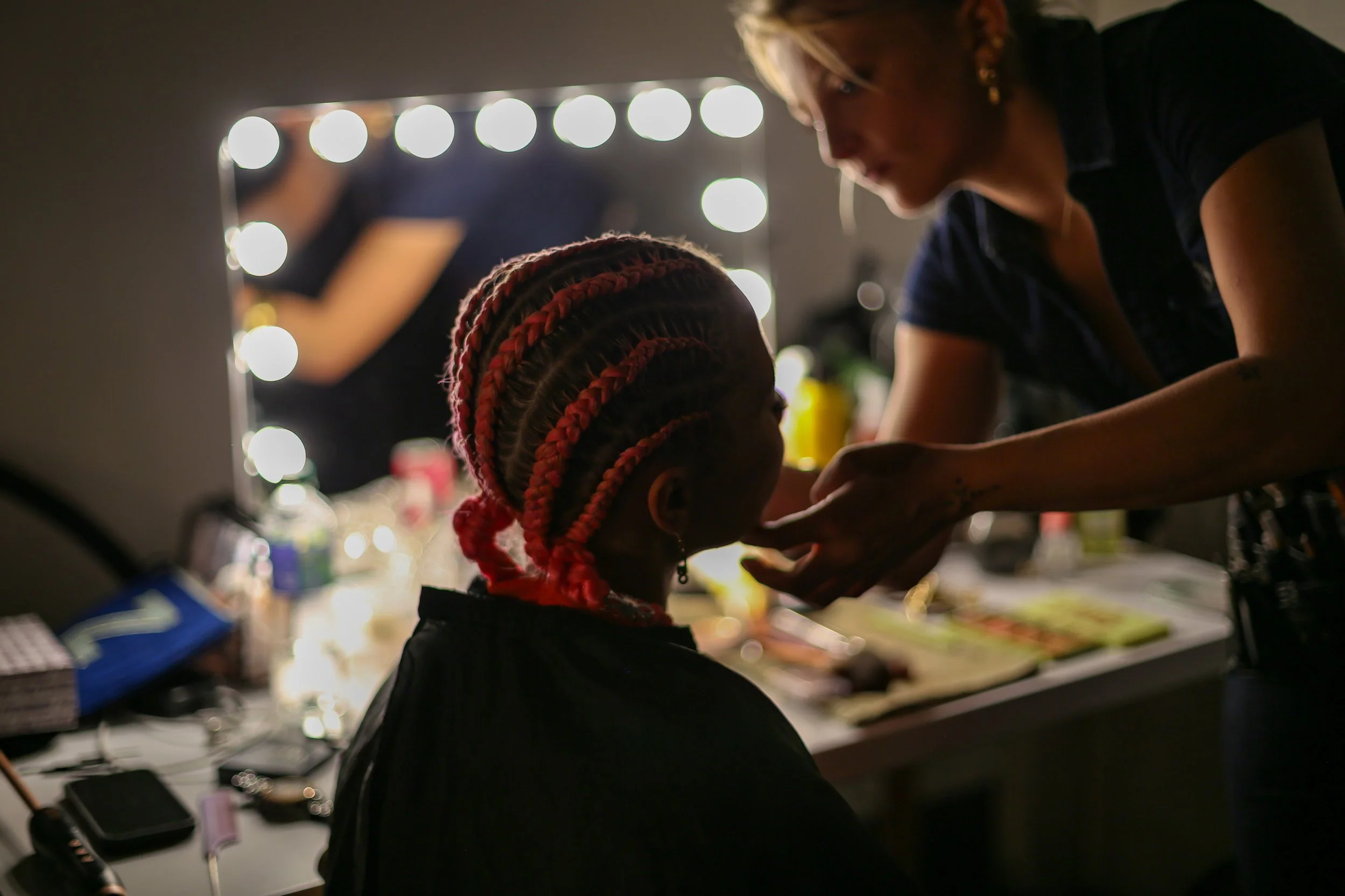 A woman with blonde hair and sunglasses leaning over a young girl with braided hair, in a well-lit workspace with a mirror and various makeup items.