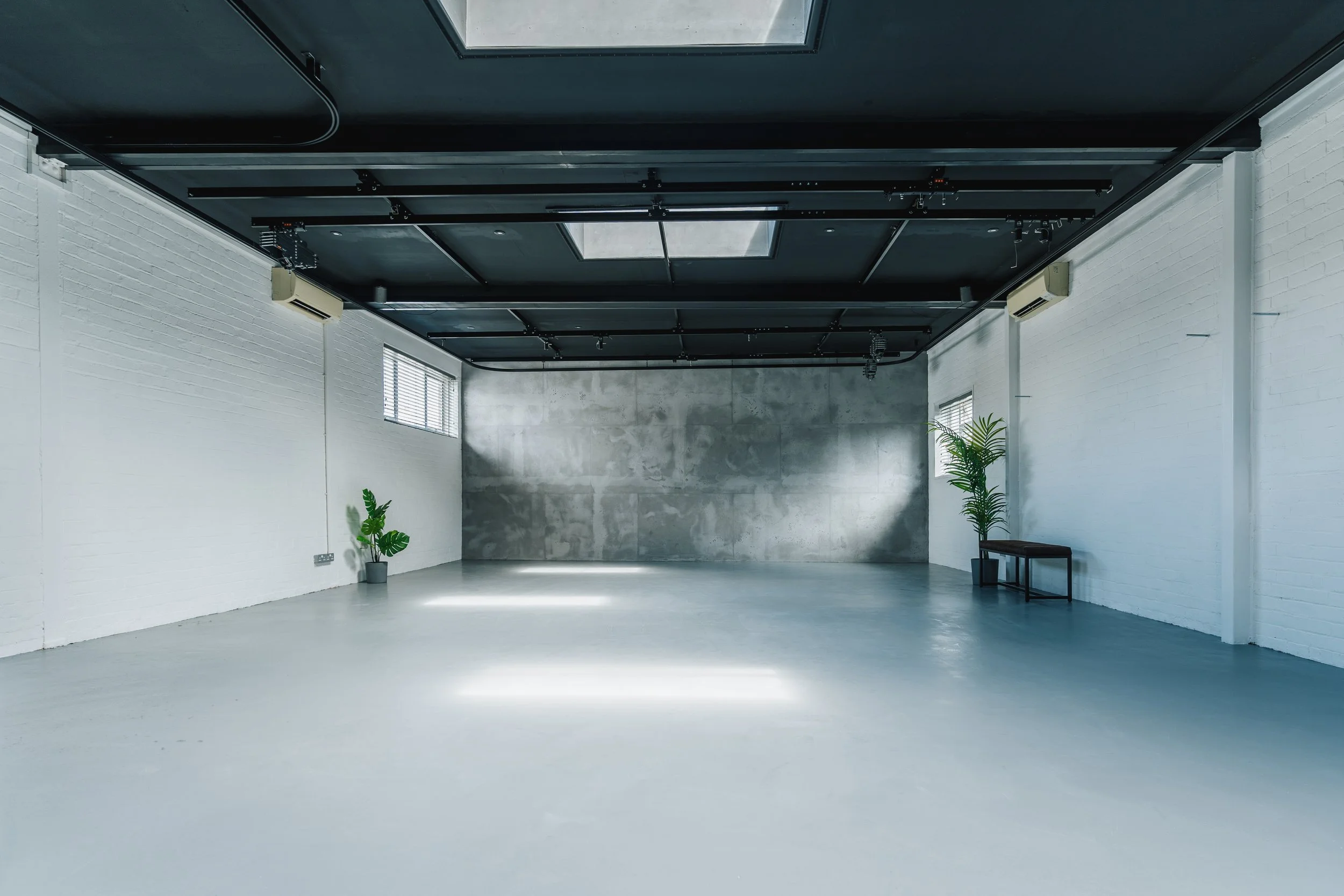 Empty room with white brick walls, a concrete accent wall, large windows with blinds, a black painted ceiling with track lighting, ceiling skylights, two potted plants, and a small bench.