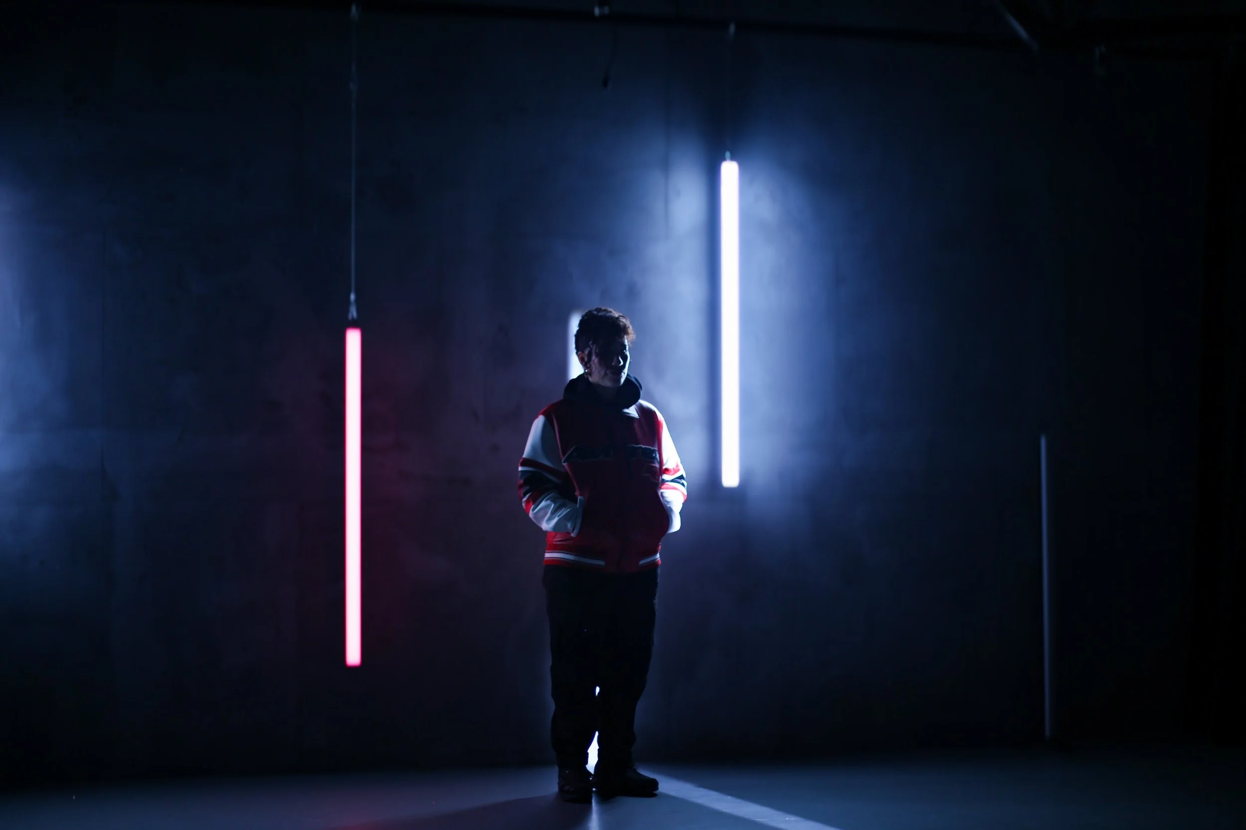 A person standing in a dark room illuminated by vertical neon lights, one pink and one white, creating a dramatic silhouette effect.