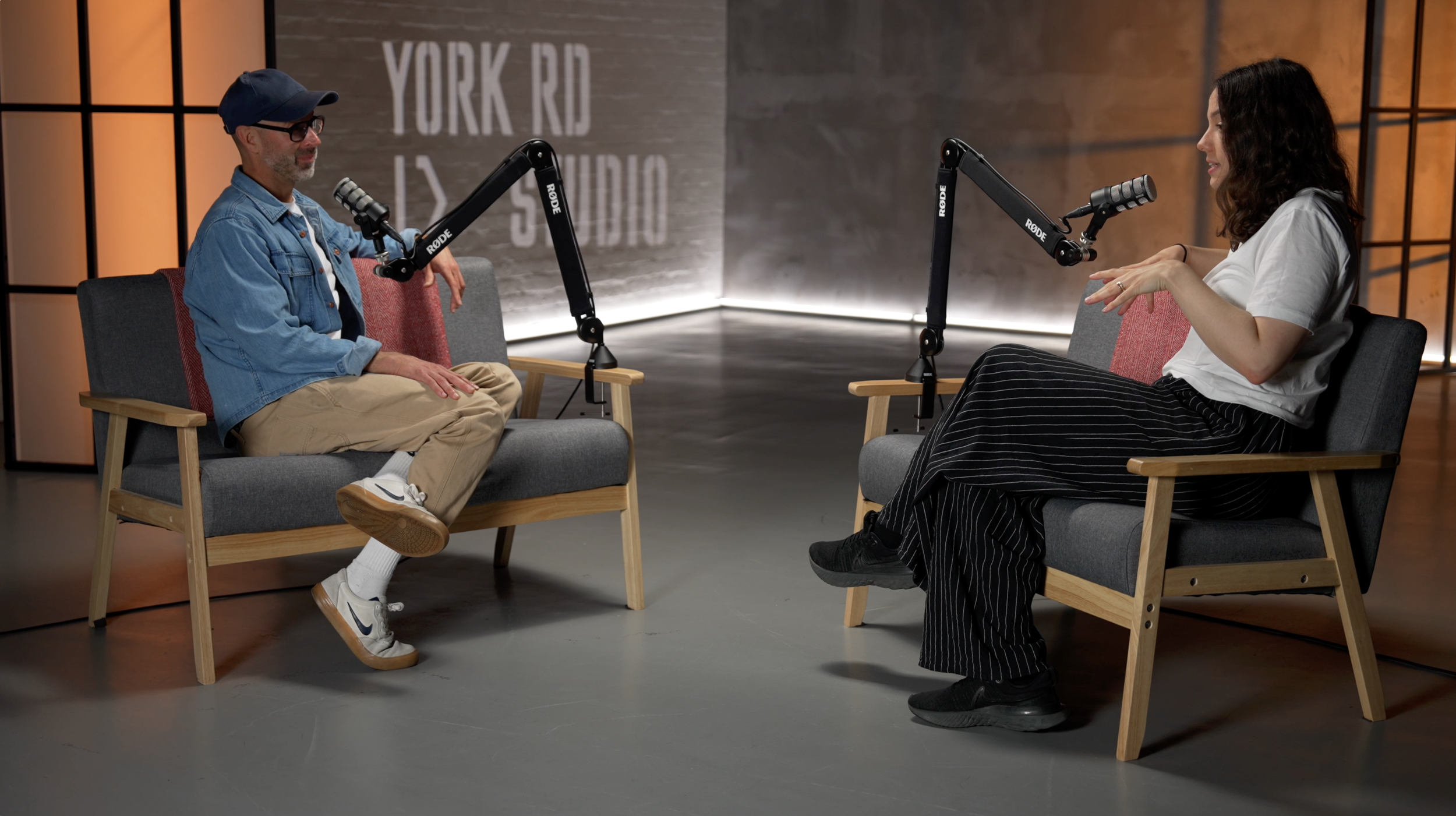 A man and woman are seated facing each other in a podcast recording studio, using microphones. The man is wearing a denim jacket, beige pants, white sneakers, glasses, and a navy cap, while the woman is dressed in a white shirt and black striped pant