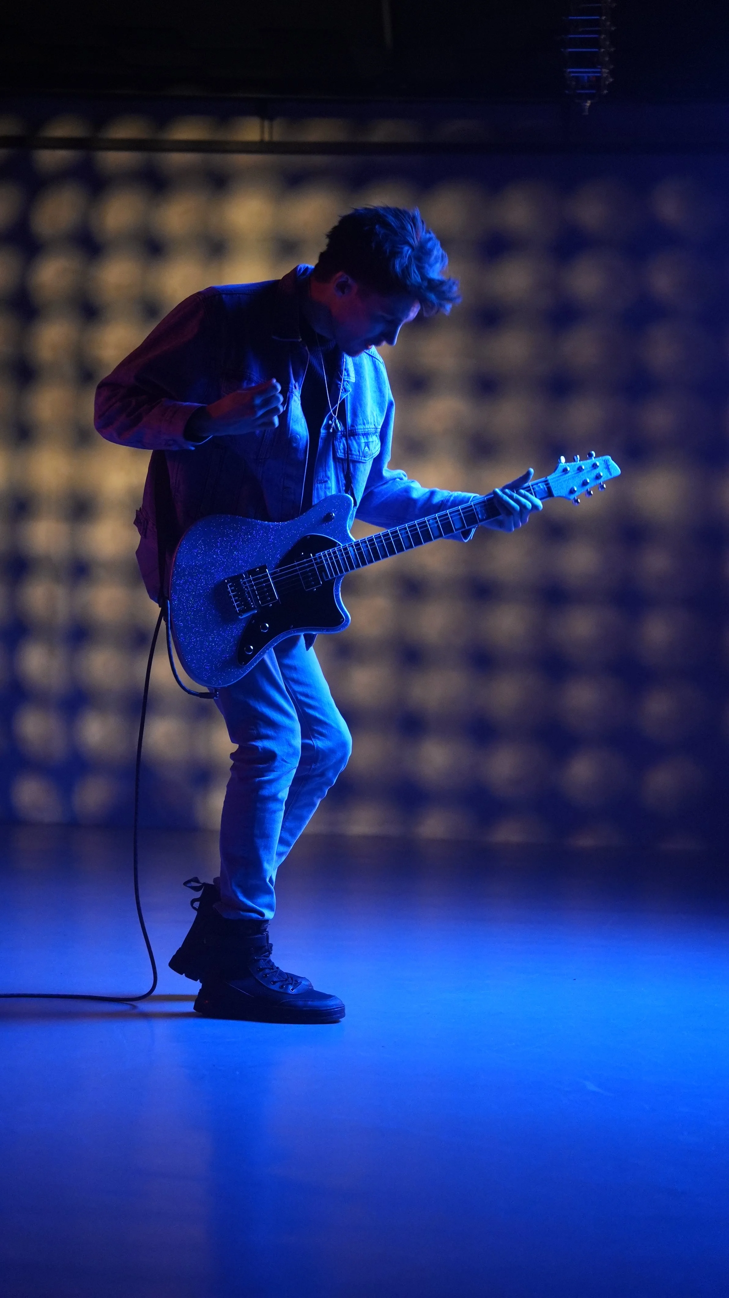 A young man playing an electric guitar on stage under blue lighting, with a blurred background and wearing casual clothes.