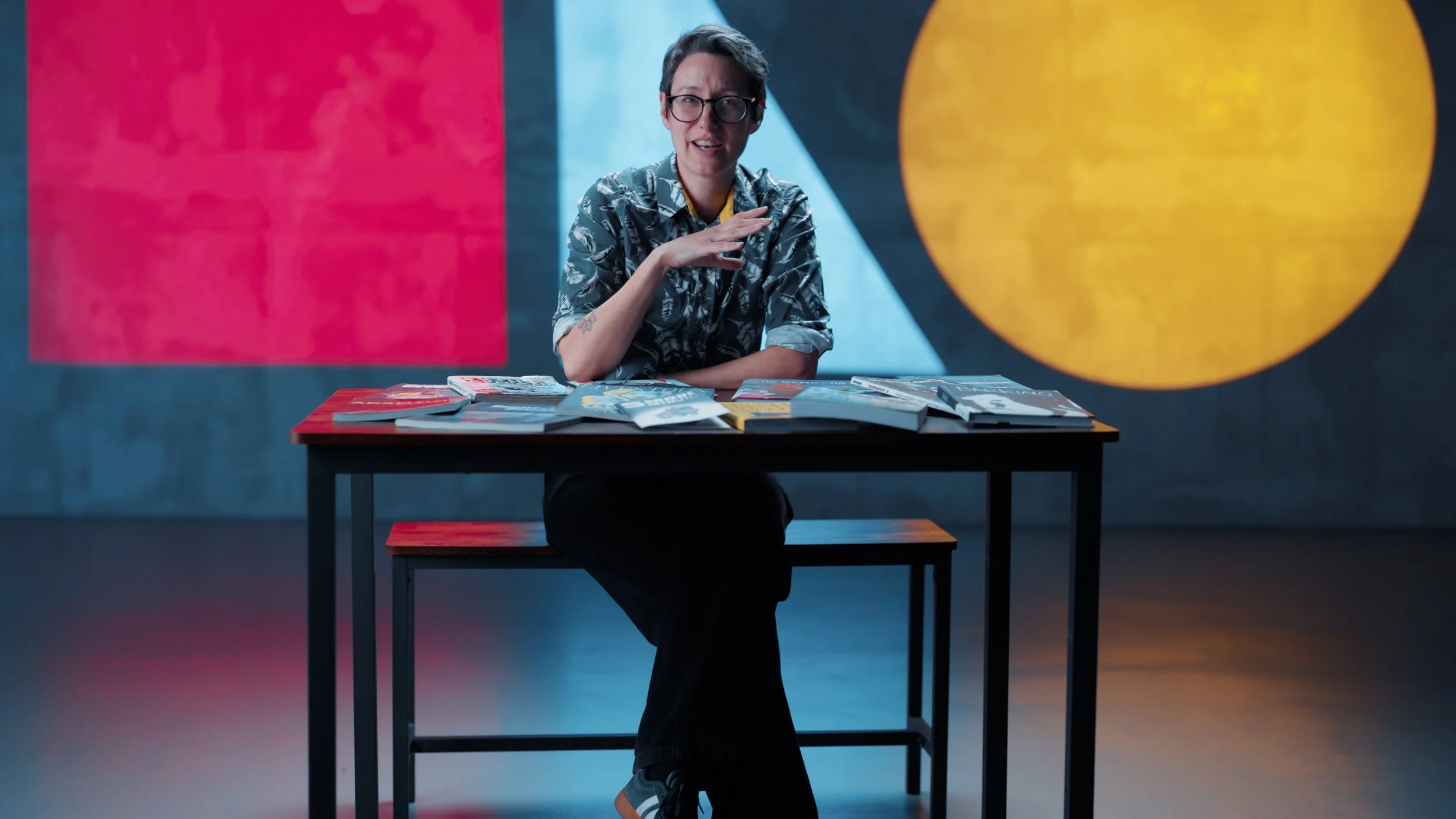 A person with short hair, glasses, and tattoos, wearing a patterned shirt, sits at a table with magazines spread out, gesturing with their right hand, in a room with colorful abstract background art.