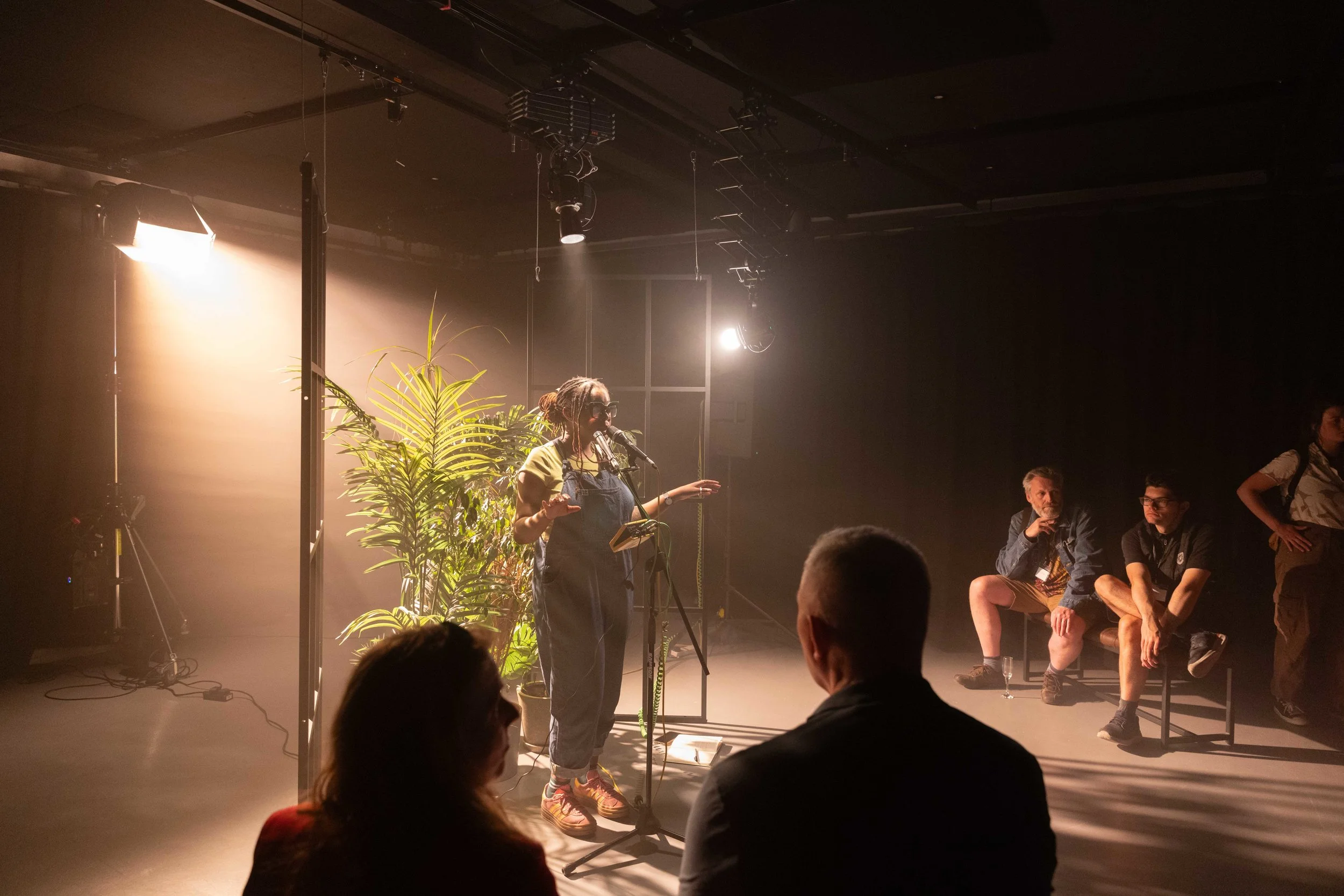 A woman wearing glasses and overalls standing in front of a plant, speaking into a microphone during a performance or presentation. Several people sit and stand nearby in a dimly lit room with stage lighting and equipment hanging from the ceiling.