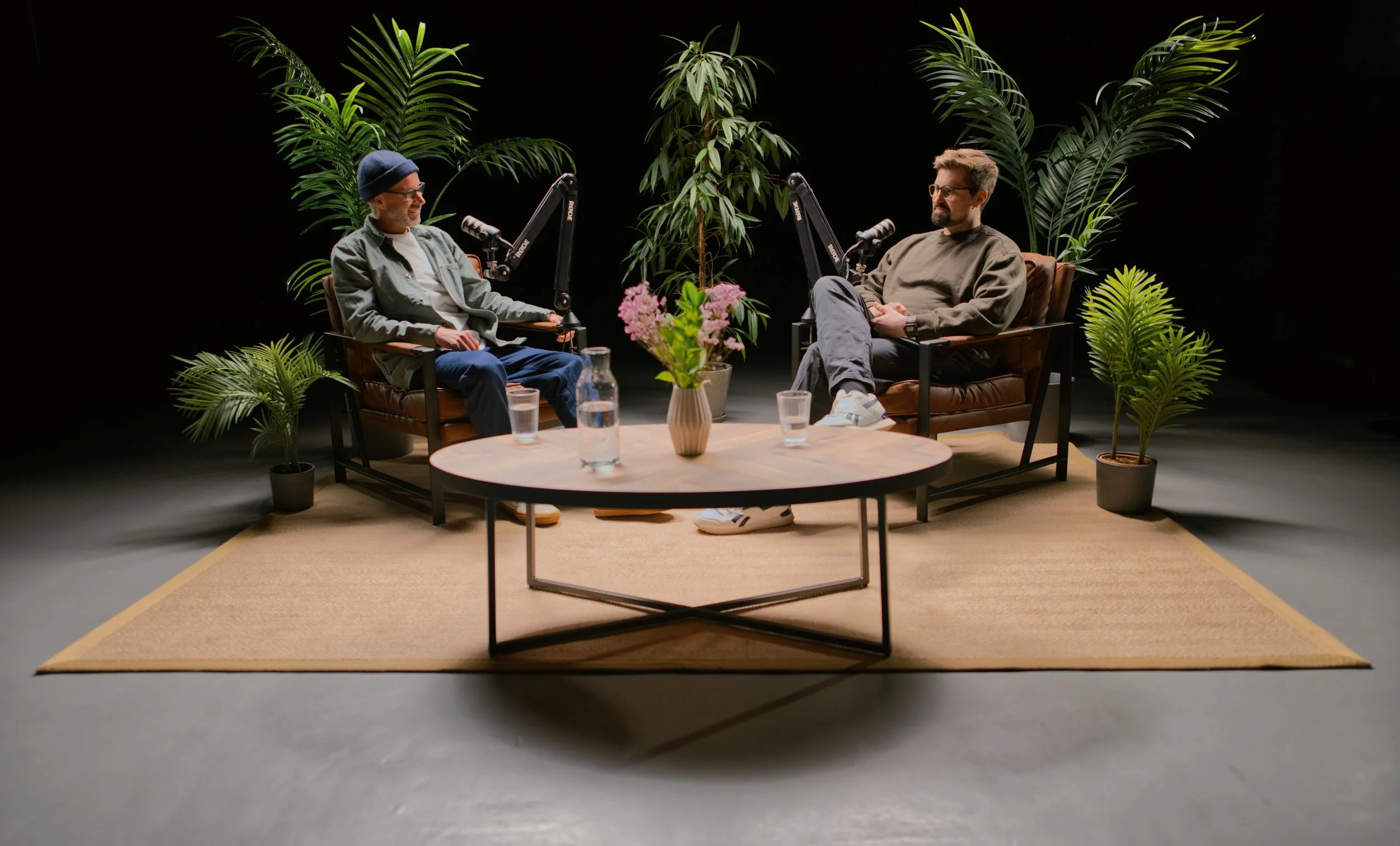 Two men sitting on chairs facing each other during a podcast or interview. They are surrounded by large green tropical plants and a round table with pink flowers and glasses of water.