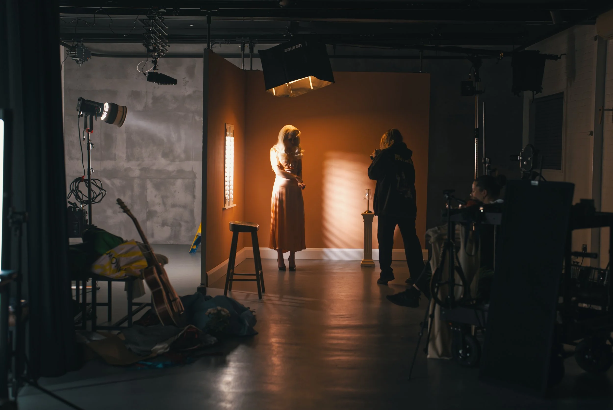 A photography studio with a woman in a beige dress standing on a pedestal, being filmed or photographed by a photographer wearing a dark hoodie. The scene is lit with professional studio lights, and there is a warm-colored backdrop behind the woman. 