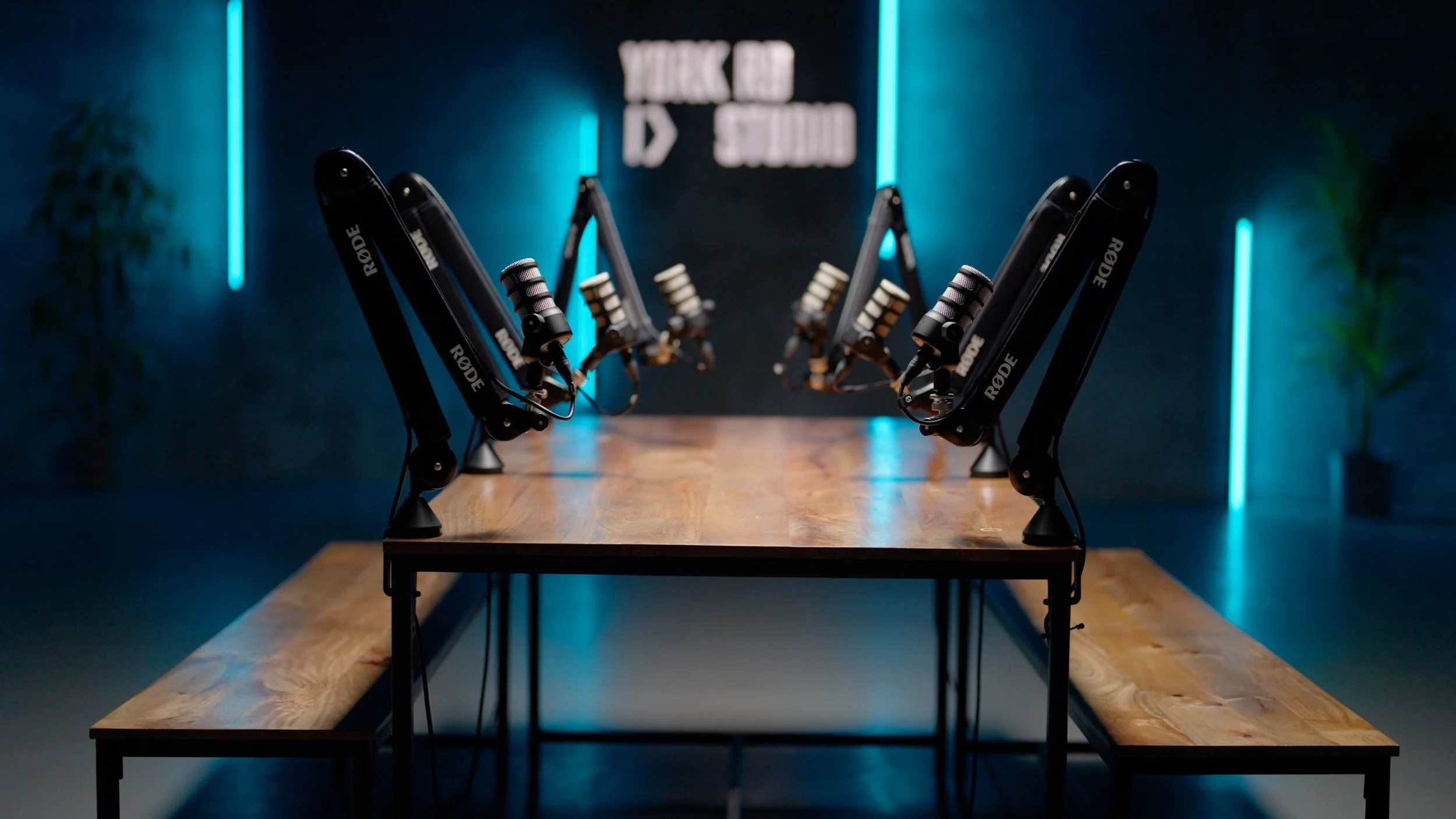 Multiple black robotic arms with mounted microphones are arranged on a long wooden table in a dimly lit room with blue neon lights, and a sign that says 'YORK STUDIOS' in the background.
