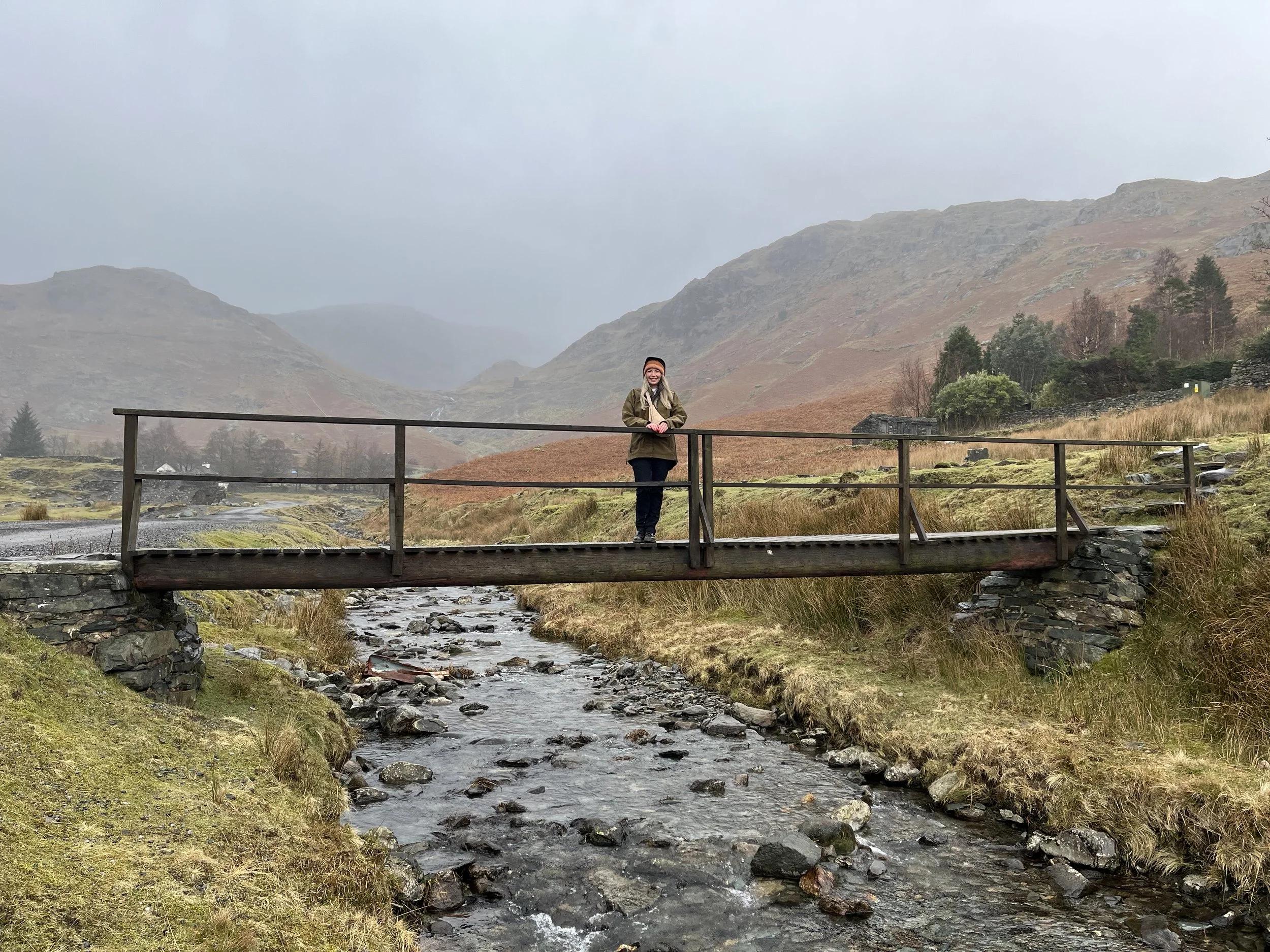 A therapist standing on a wooden bridge over a small rocky stream in a mountainous area during overcast weather.