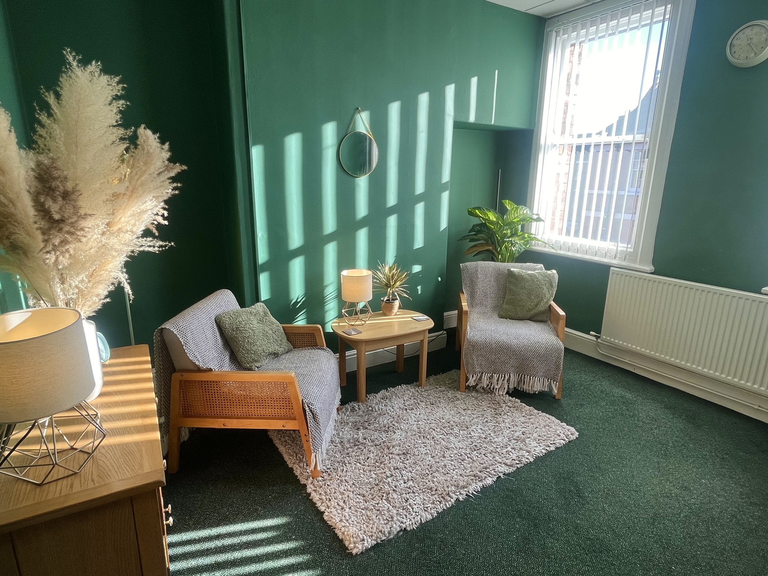 A cozy therapy room with green walls, two gray armchairs with wooden frames for counselling sessions, a small wooden side table with a lamp, a fluffy beige rug, large window with blinds, potted plants, and a round wall mirror.