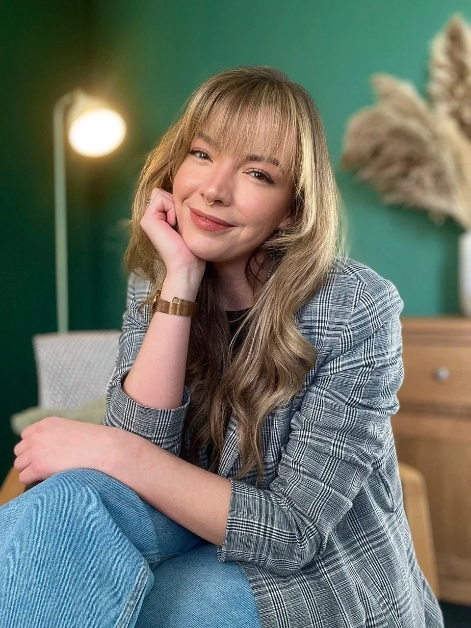 A psychotherapeutic counsellor with blonde hair, smiling softly, resting her chin on her hand, sitting in a therapy room with a green wall, a lamp, and decorative dried plants in the background.