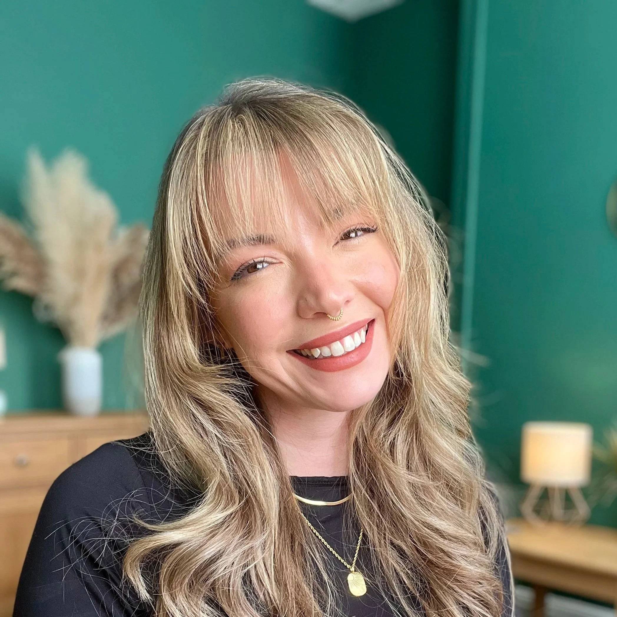 A smiling psychotherapeutic counsellor with wavy blonde hair and a septum piercing, wearing a black top and layered gold necklaces, in a therapy room with a green wall and a blurred background.
