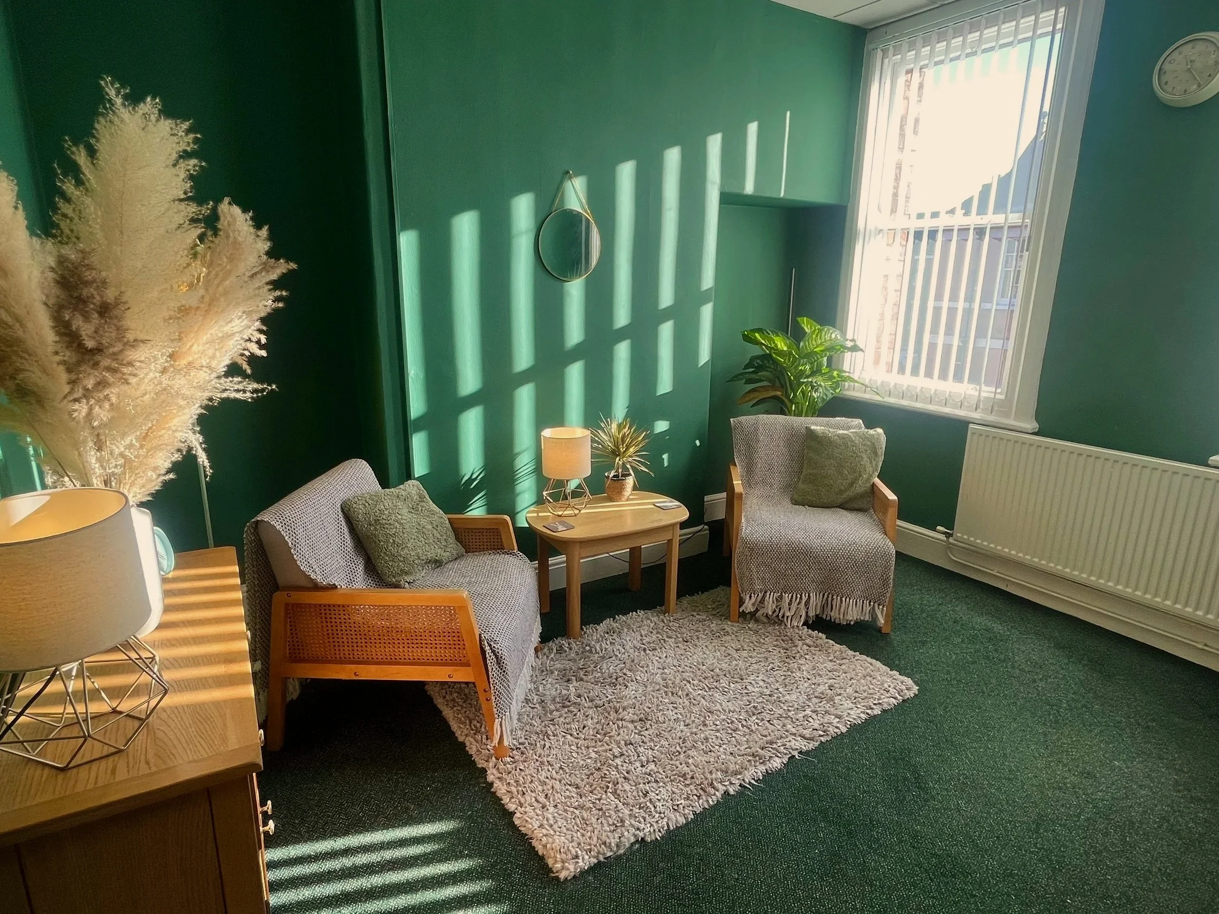 Counselling room in Nantwich with green walls, two armchairs with throws and pillows, a wooden side table with a lamp, a shaggy rug, a large window with blinds, potted plants, a small mirror on the wall, and a clock.