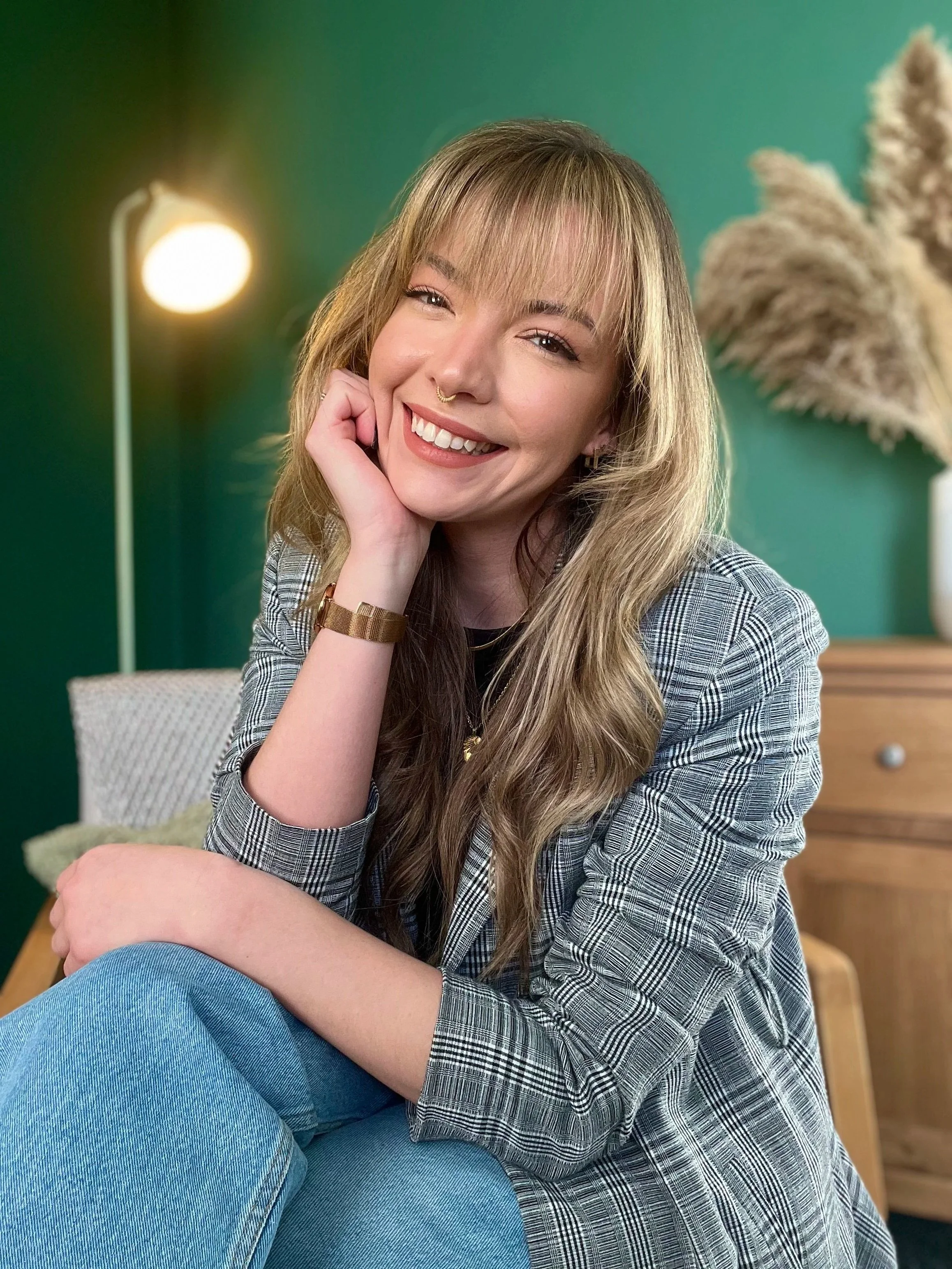 A smiling therapist with blonde hair and a septum piercing, wearing a gray plaid blazer and jeans, sitting indoors in front of a green wall in a therapy room with a lamp and decorative plants.