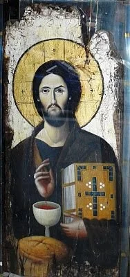 A religious icon painting of Jesus Christ holding a bread and a cup, with a halo around his head, set against a weathered, aged background.