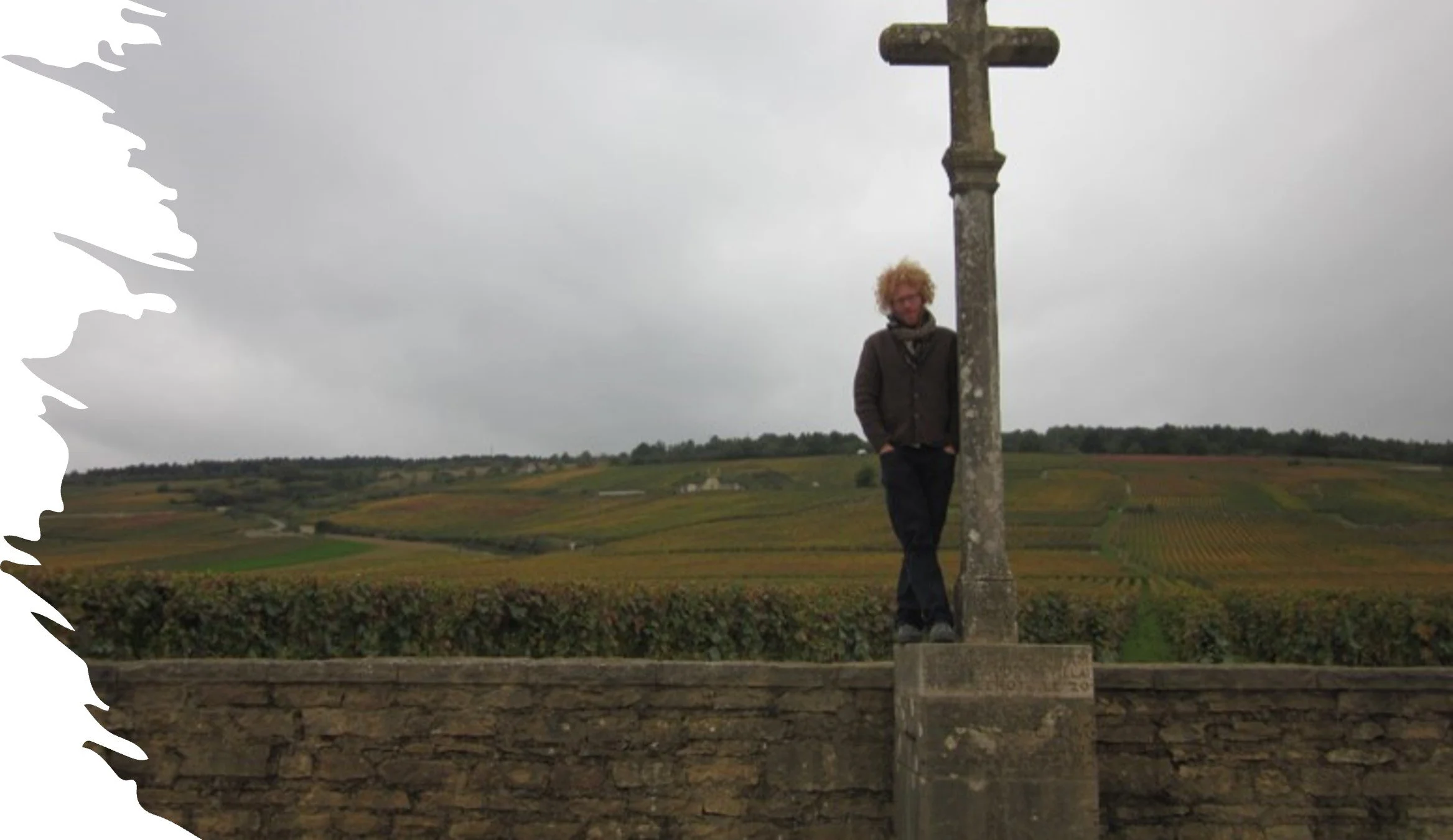 A person with curly blonde hair standing next to a large stone cross in a rural landscape with rolling fields under a cloudy sky.