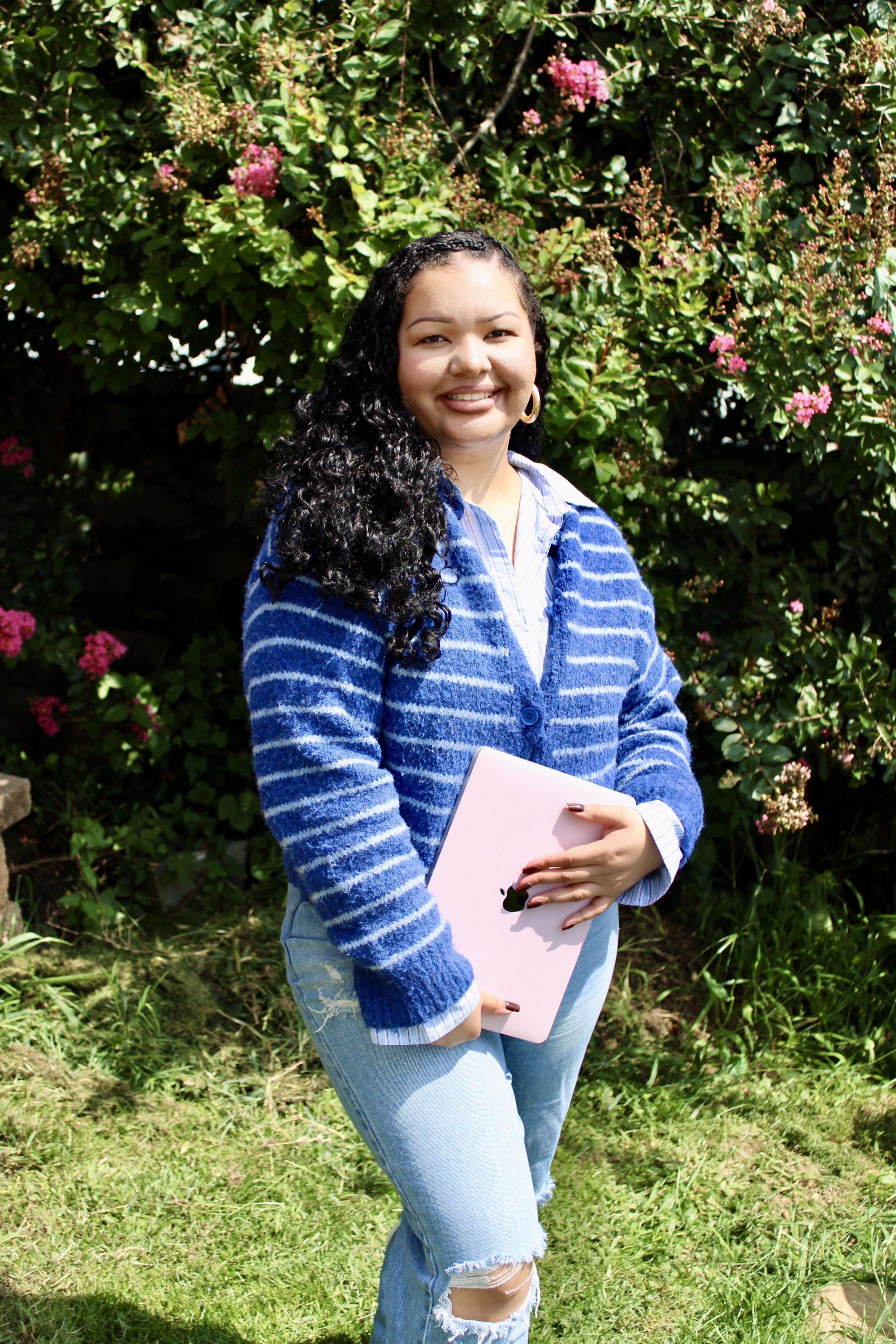 A woman with long, curly black hair, wearing a blue and white striped cardigan, light blue ripped jeans, and hoop earrings, holding a pink notebook, standing outdoors in front of green foliage with pink flowers.