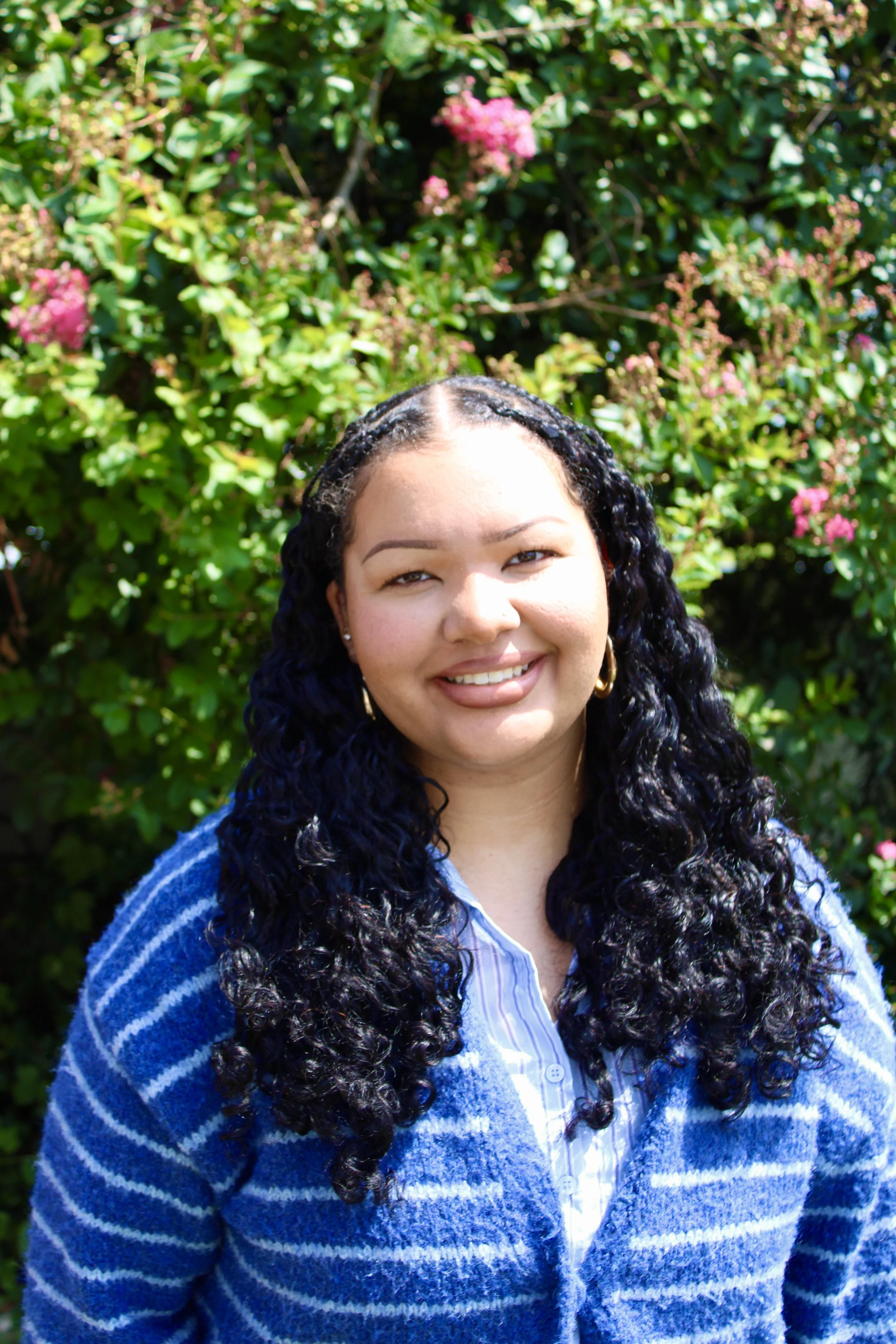 A woman with curly black hair and earrings smiling outdoors, wearing a blue and white striped sweater and blouse, standing in front of green foliage with pink flowers.