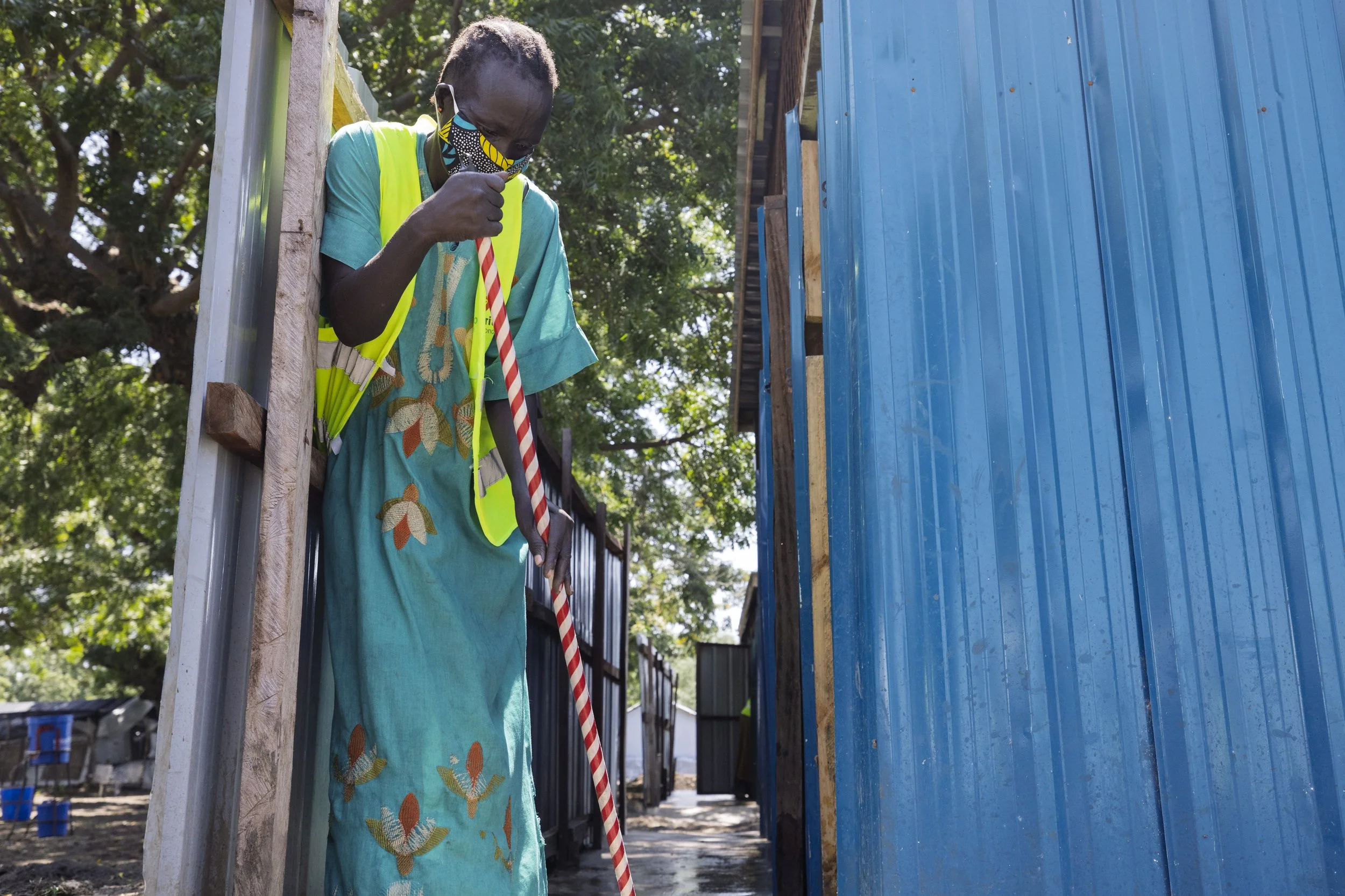 A person wearing a colorful traditional dress, a yellow safety vest, and a patterned face mask, standing outdoors between trees and blue structures, holding a striped cane.
