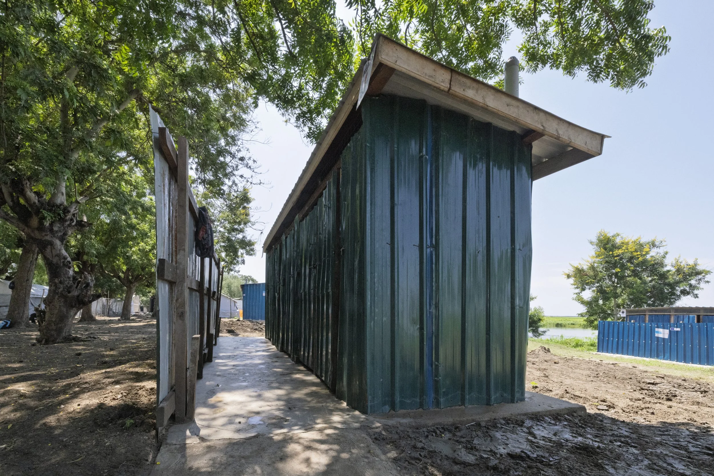 A small, green metal shed with a sloped roof, situated outdoors on a concrete slab, surrounded by trees and dirt ground, with blue fencing in the background.