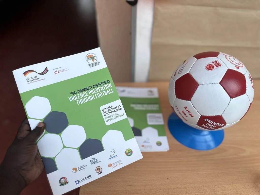 Two people are working in a modern office, with a large monitor and two computers on the desk. The woman is using a keyboard and mouse, while the man is holding a notebook and speaking. There are screens and camera equipment in the background, and a large screen on the wall displaying a person's face.