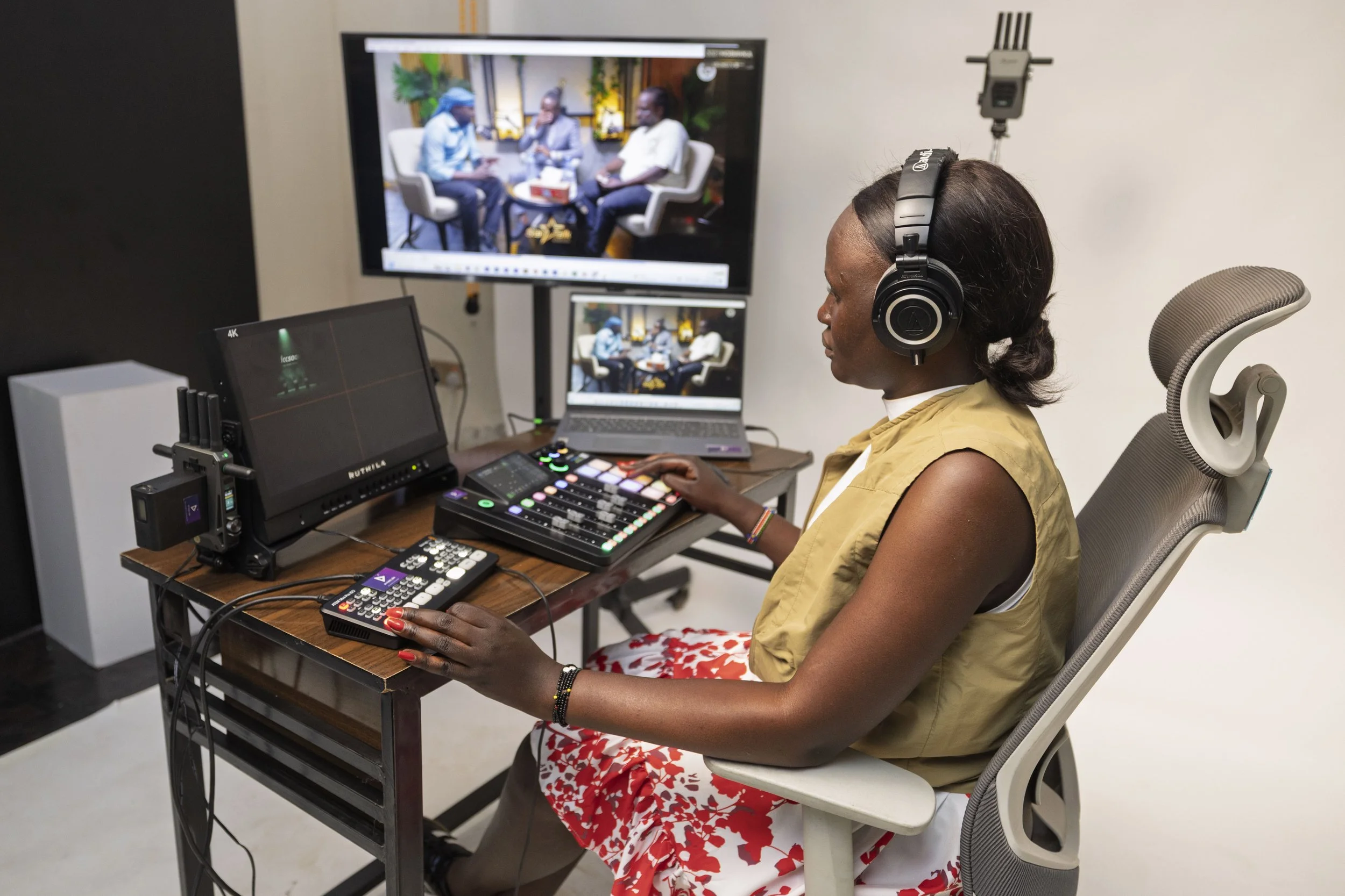 A woman in a beige vest and red floral skirt sits at a desk wearing large headphones and operating video editing equipment. She is in a studio with multiple monitors displaying the same image of a group of people in a talk show setup.