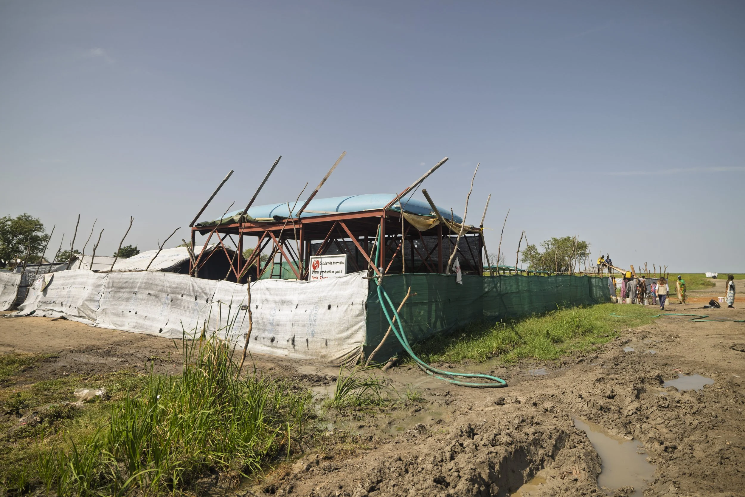 Temporary water production thatched structure with a sign, fencing, and people nearby in a rural area.