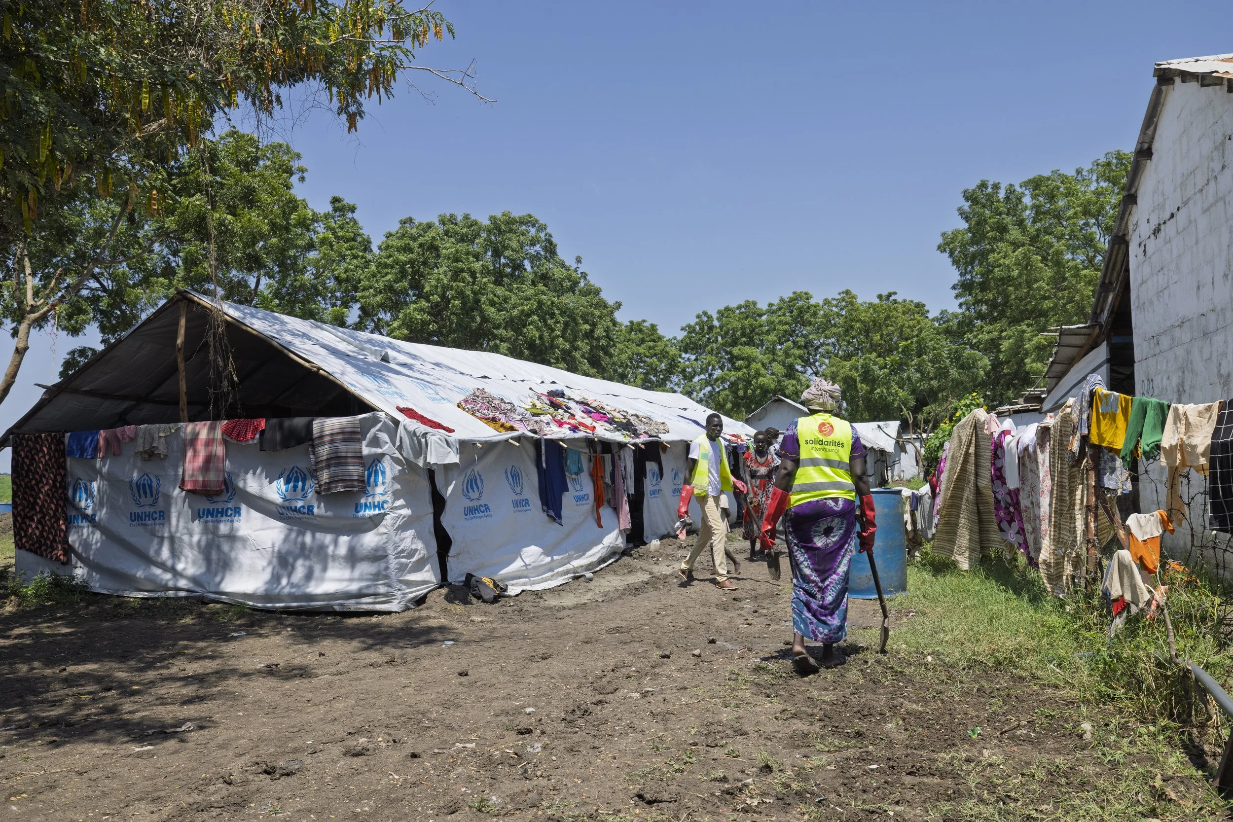 People walking through a refugee camp with tents and clothes hanging to dry, surrounded by trees under a clear blue sky.