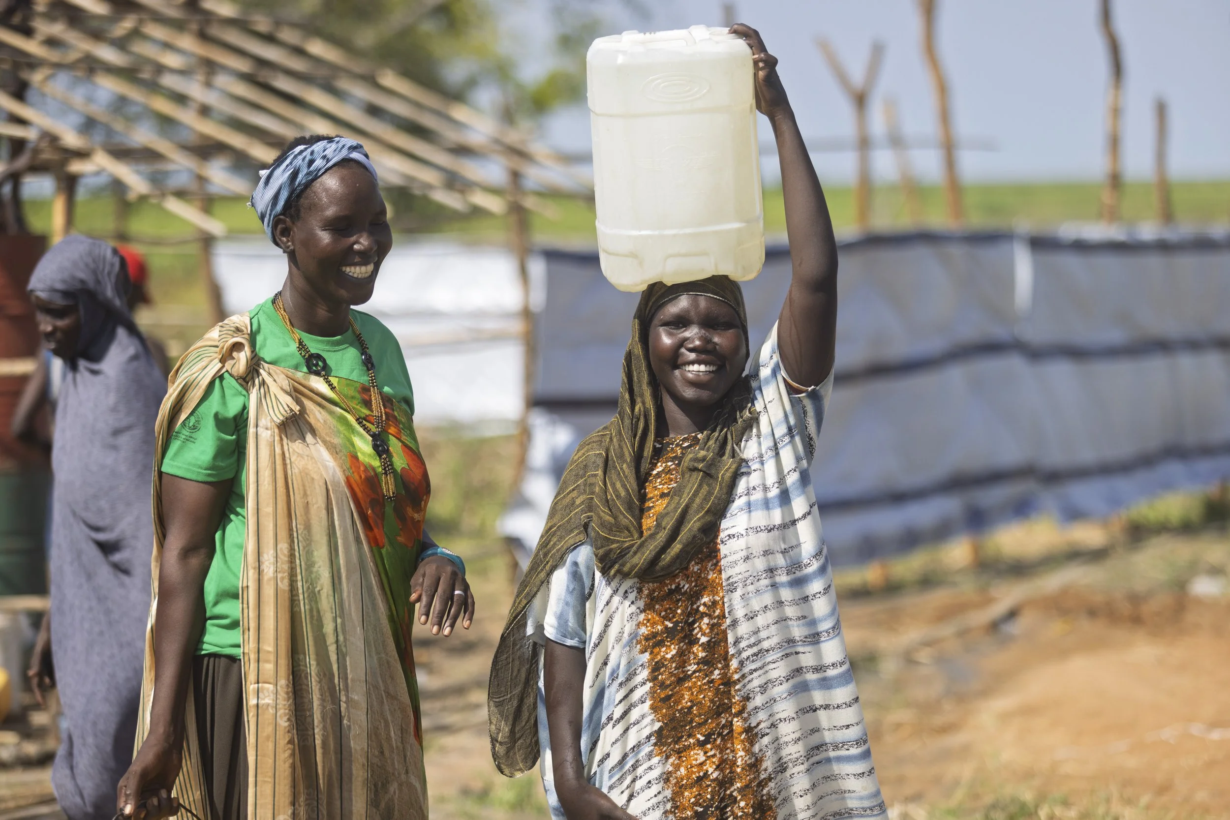 Two smiling women, one carrying a plastic container on her head in an outdoor rural setting, with structures and greenery in the background.