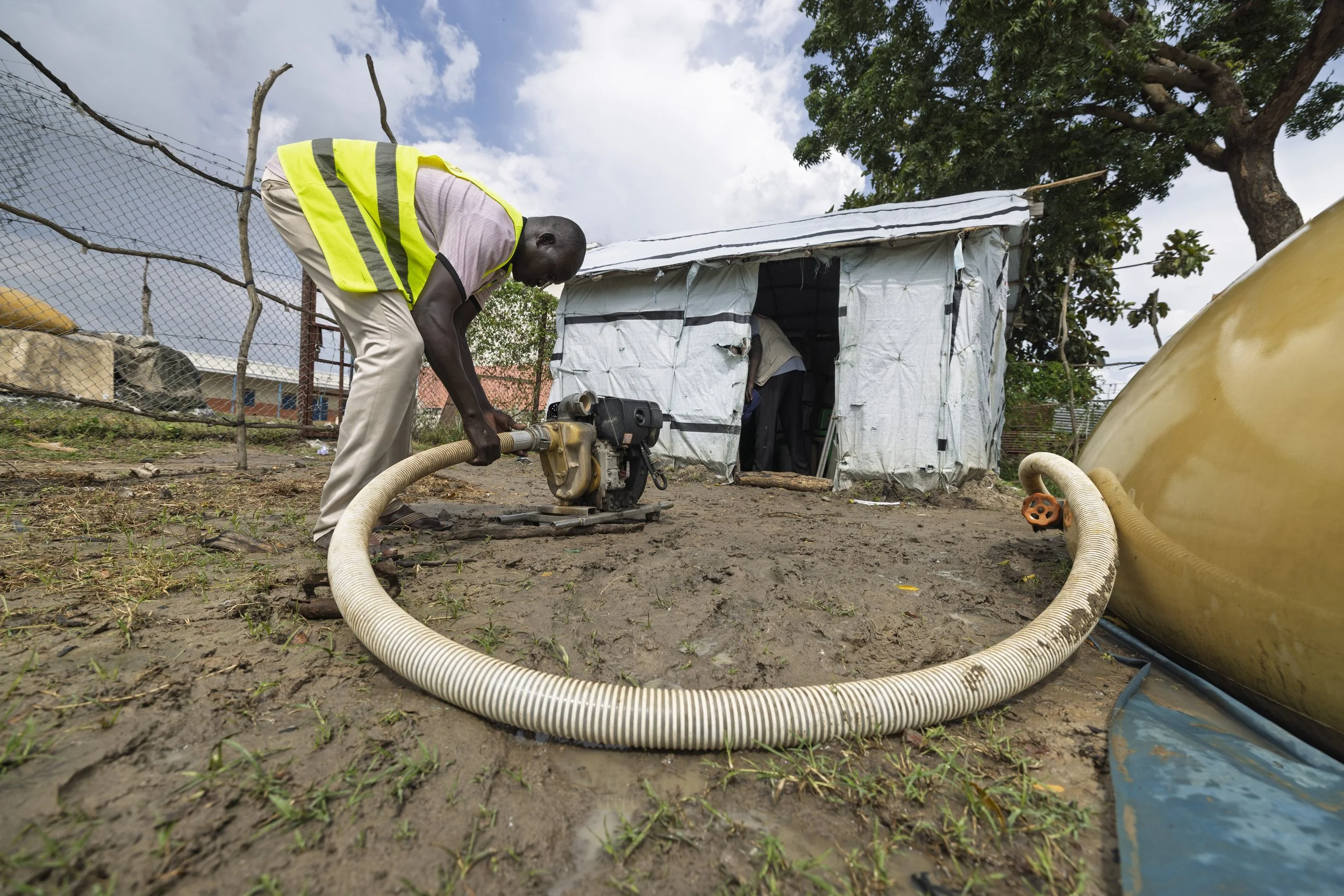A man wearing a yellow safety vest is operating a generator in front of a small, makeshift shelter made of white tarp. The ground appears muddy, and there is a large yellow tank on the right side of the image. A fence and trees are visible in the bac