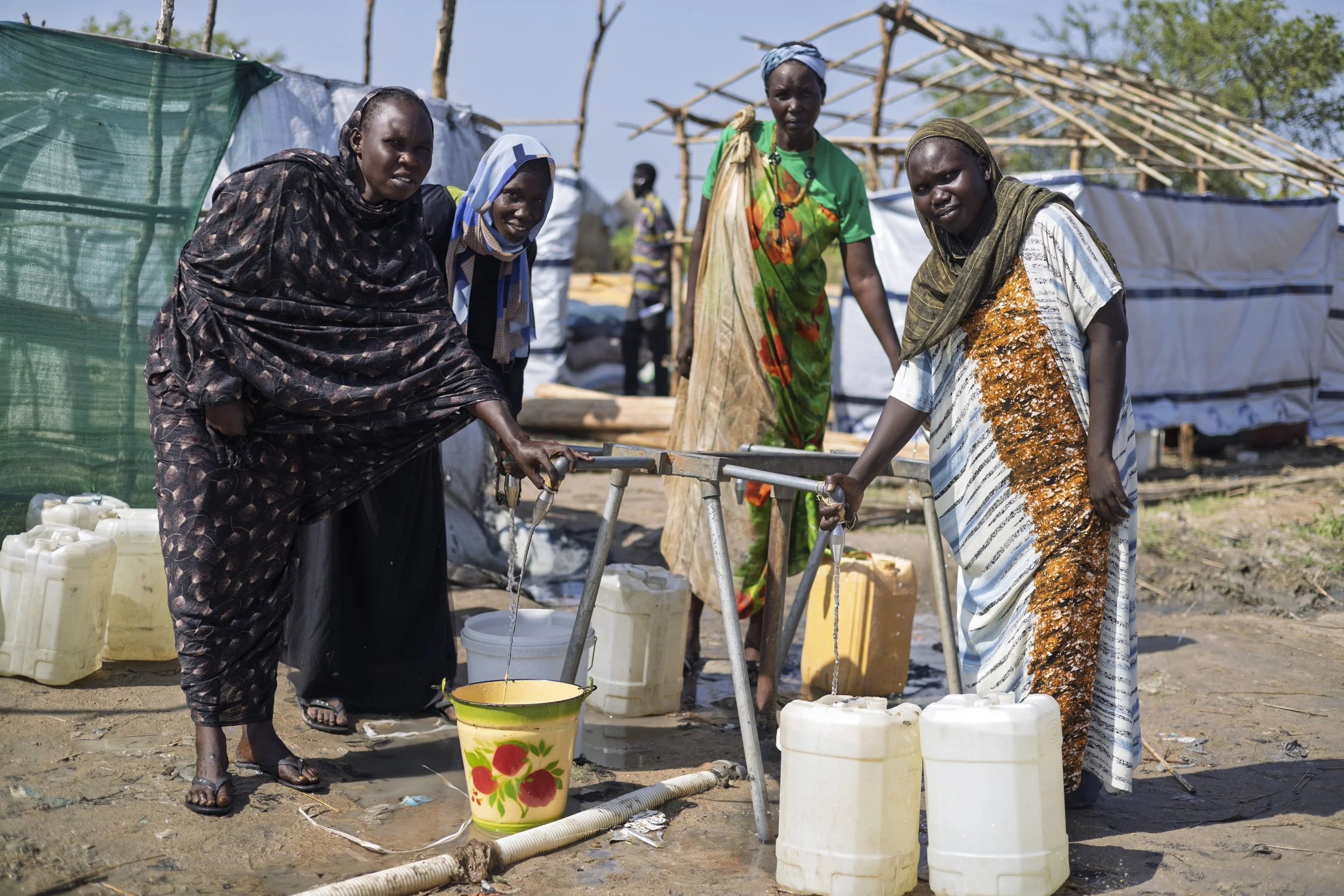 Four women in colorful dresses filling containers with water from a hand pump in a refugee camp with temporary shelters in the background.