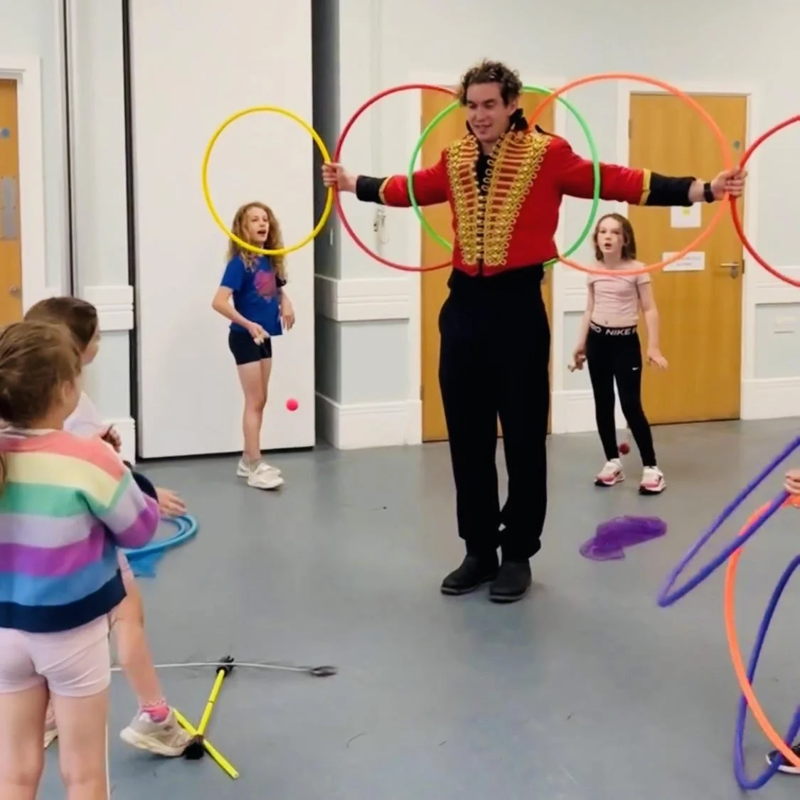 A performer dressed as a circus ringmaster standing with arms outstretched, holding multiple hula hoops in a room with children watching. Three girls are nearby, one holding a hula hoop and two in the background with a smile.