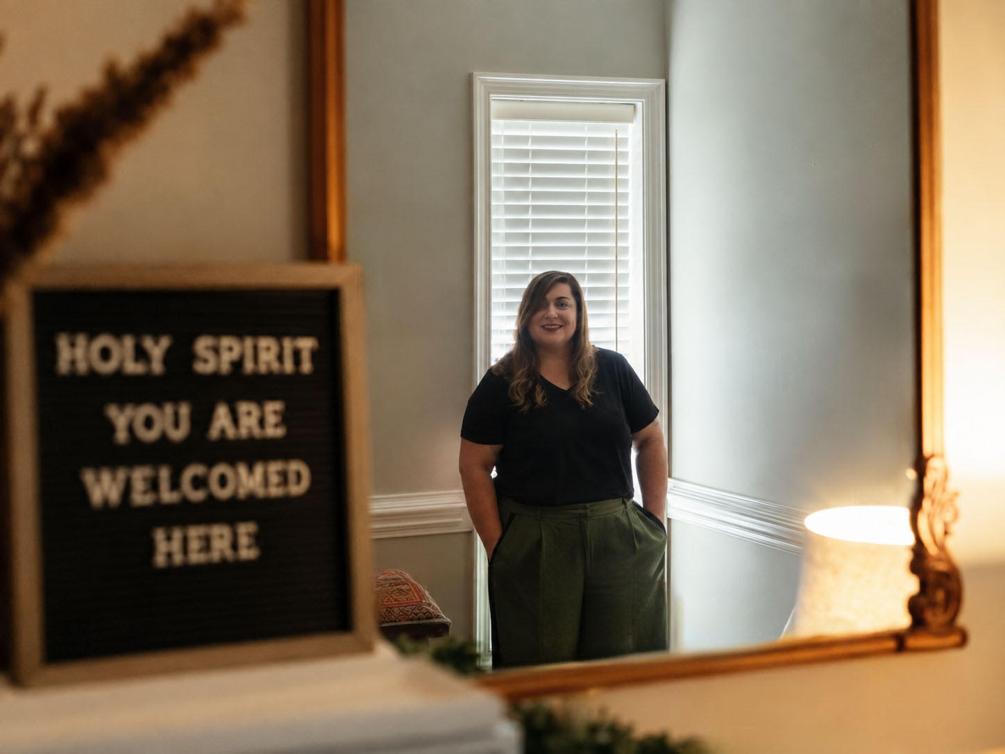 Steffany Medina reflected in a gold-framed mirror beside a sign welcoming the Holy Spirit.