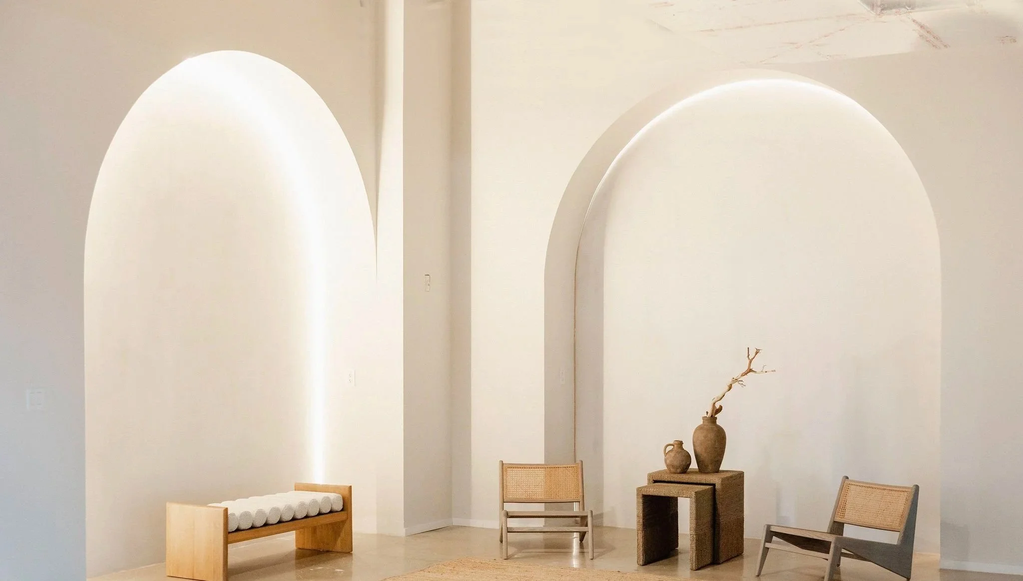 Minimalist interior with white walls and arched wall niches. There are three wooden chairs, two with woven seats, and a small table holding two vases and a decorative branch. A soft beige rug is on the floor.