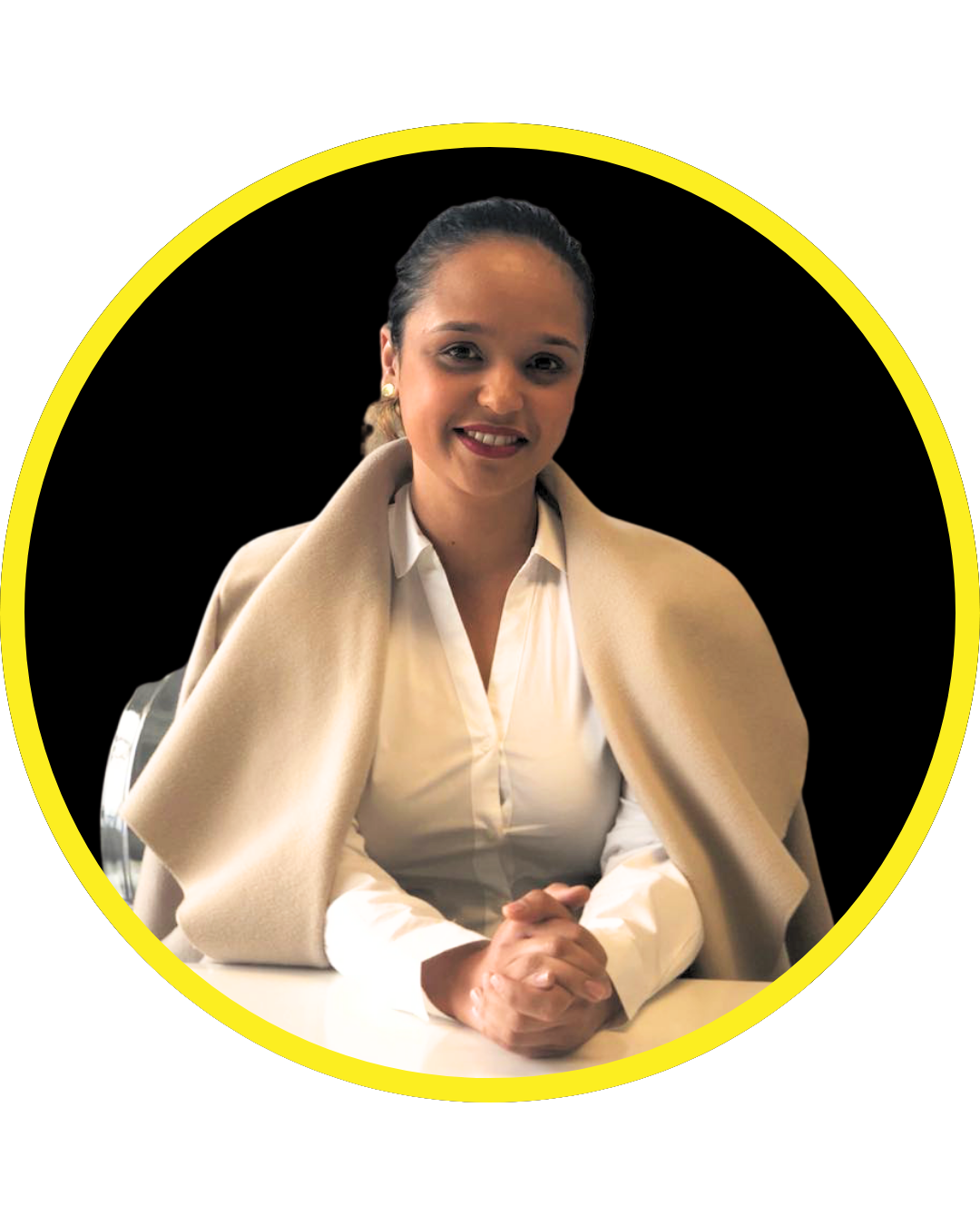 A woman with dark hair tied back, wearing a white blouse and a beige coat draped over her shoulders, smiling at the camera. She is seated at a table with her hands clasped.