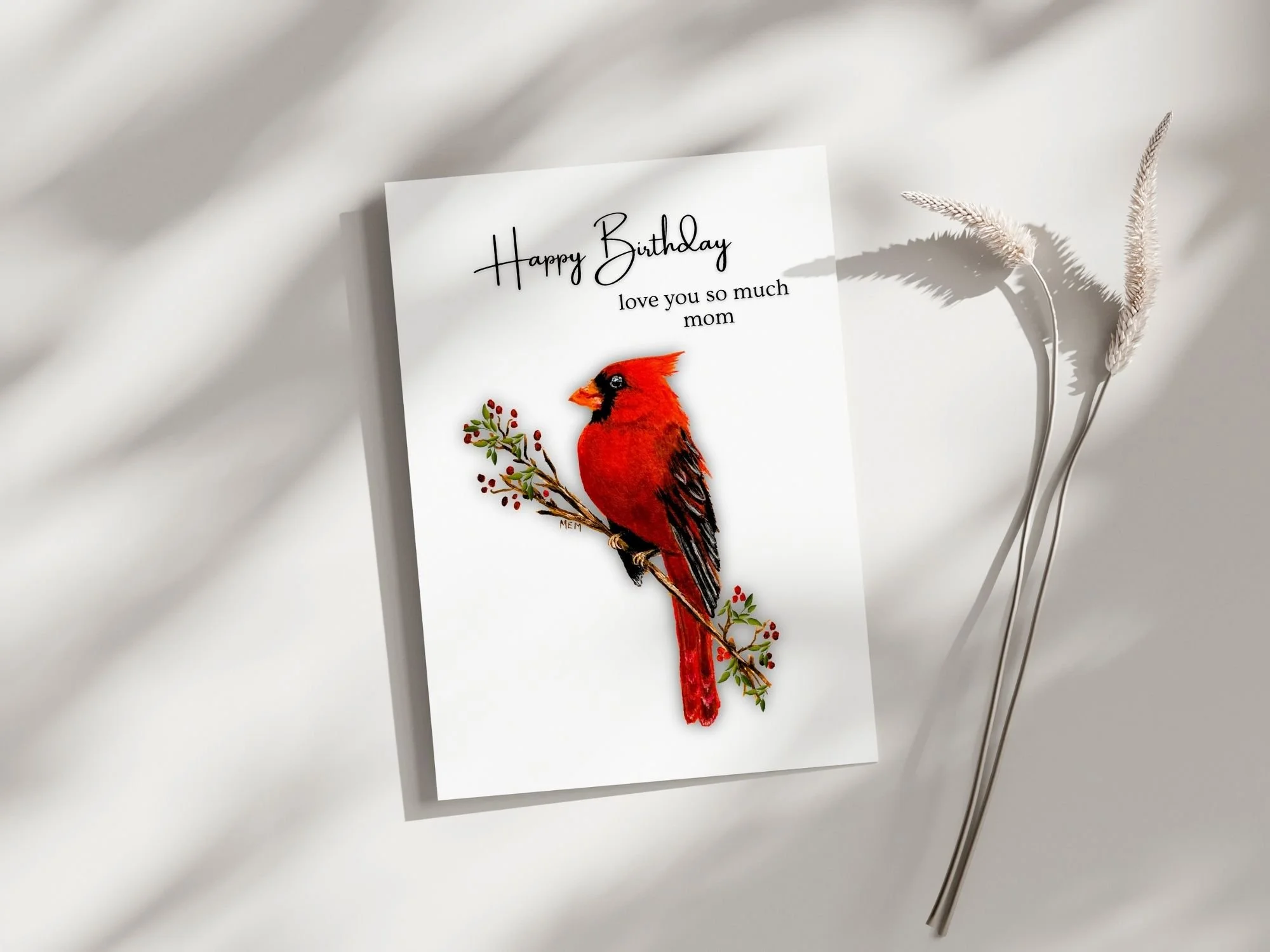 a birthday card with a red cardinal perched on a branch facing left with the words Happy Birthday love you so much mom above it.  The background is white.  Art by Maureen MacKenzie