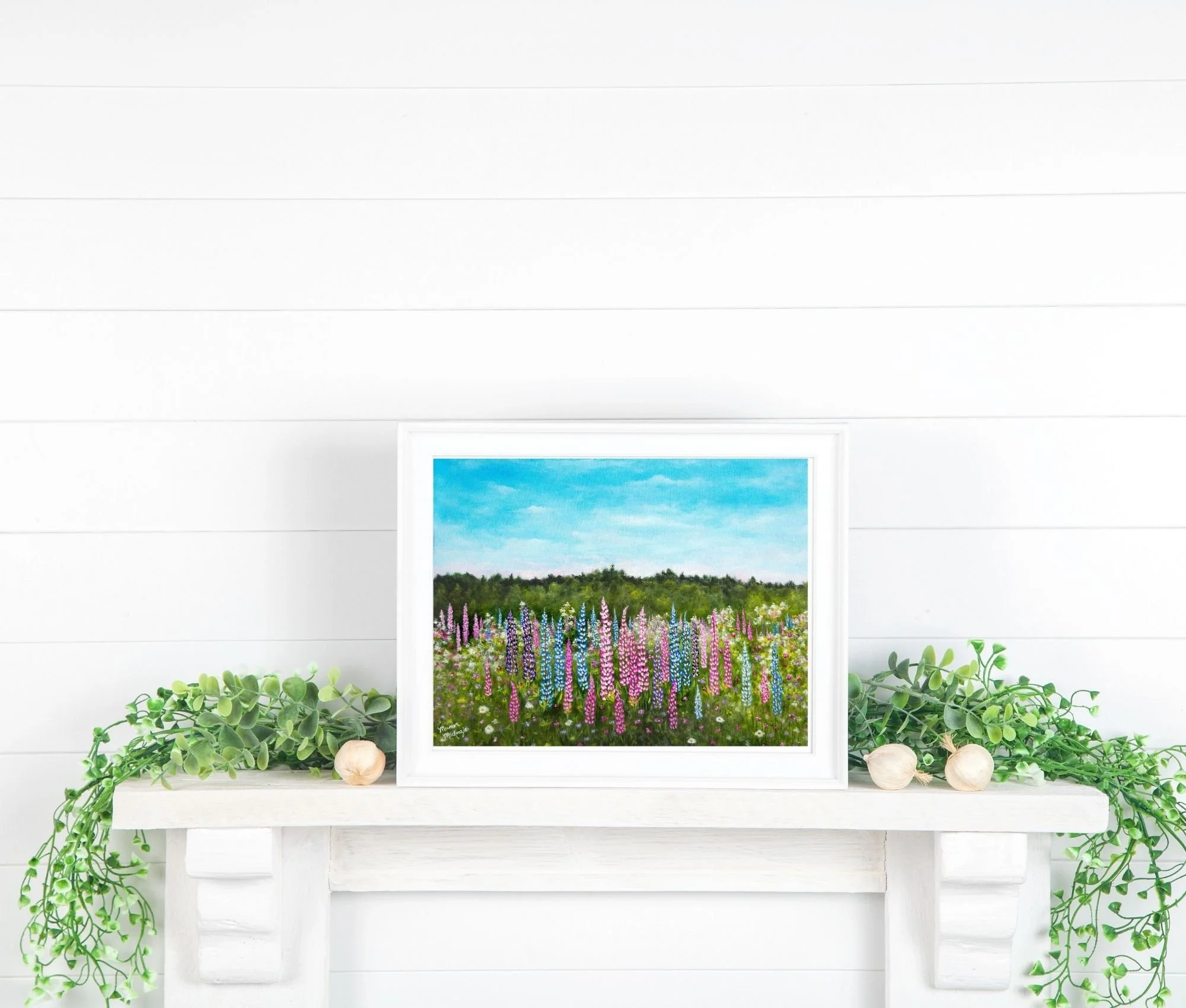 a framed print on a mantle of a colorful field of pink and purple lupines in bloom under a pale blue sky,