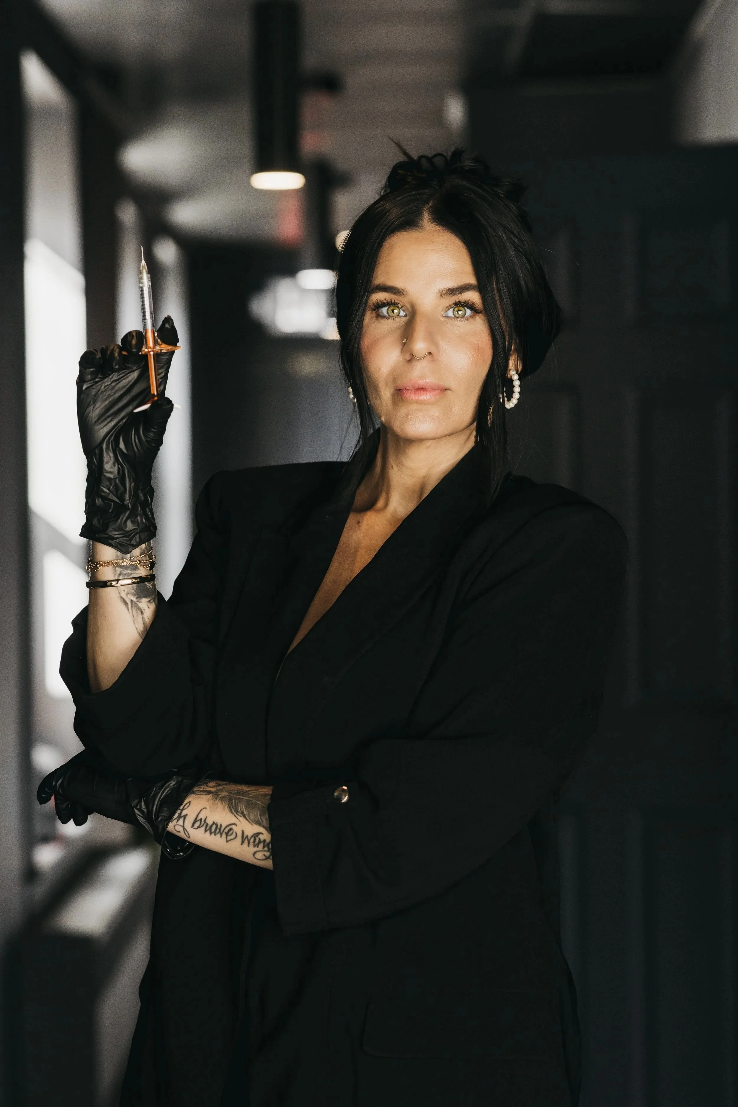 A woman with dark hair holding a syringe in a professional tattoo or medical setting, wearing black gloves and a black outfit.