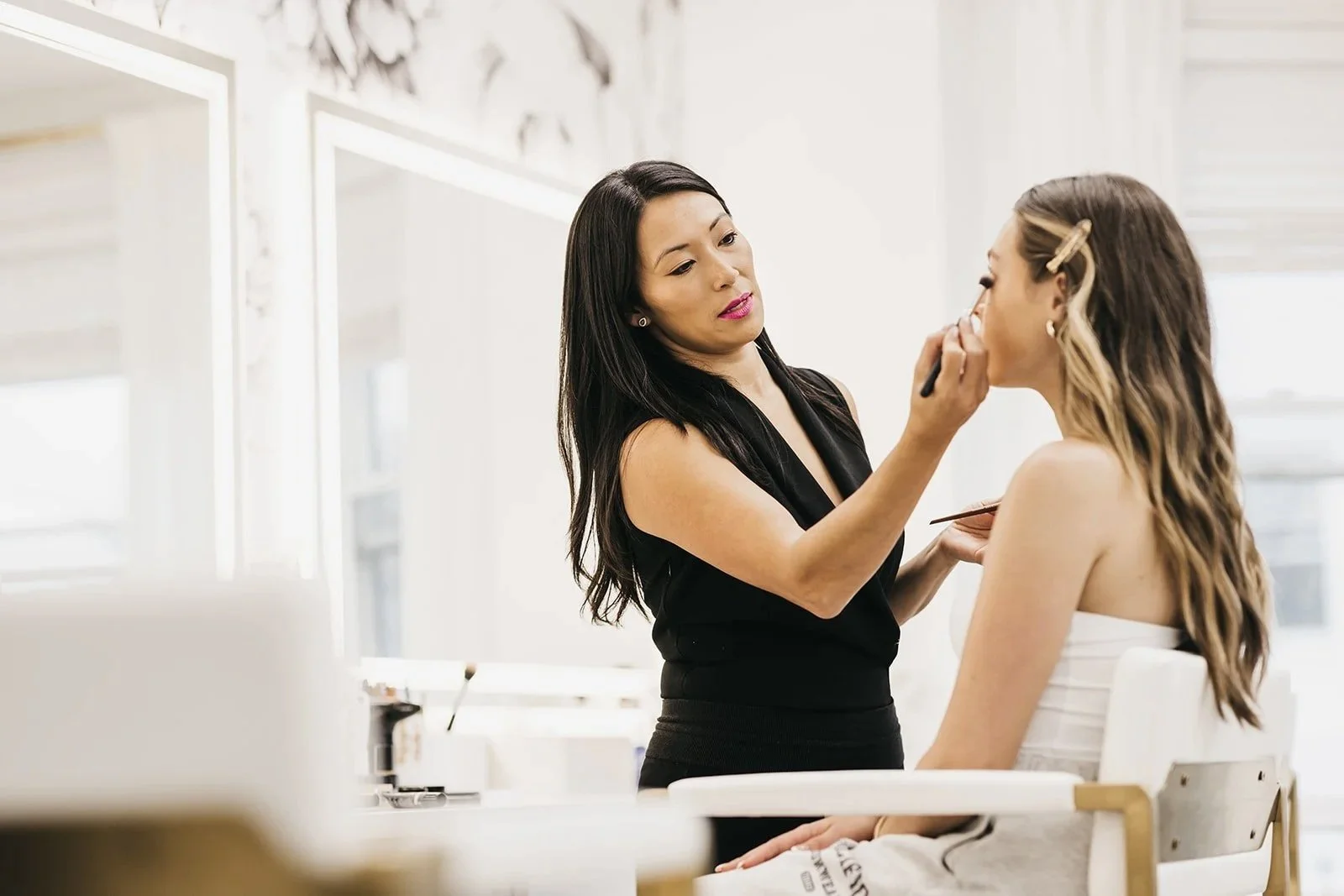 Makeup artist applying makeup to a woman seated in a salon chair, with a bright, modern interior.