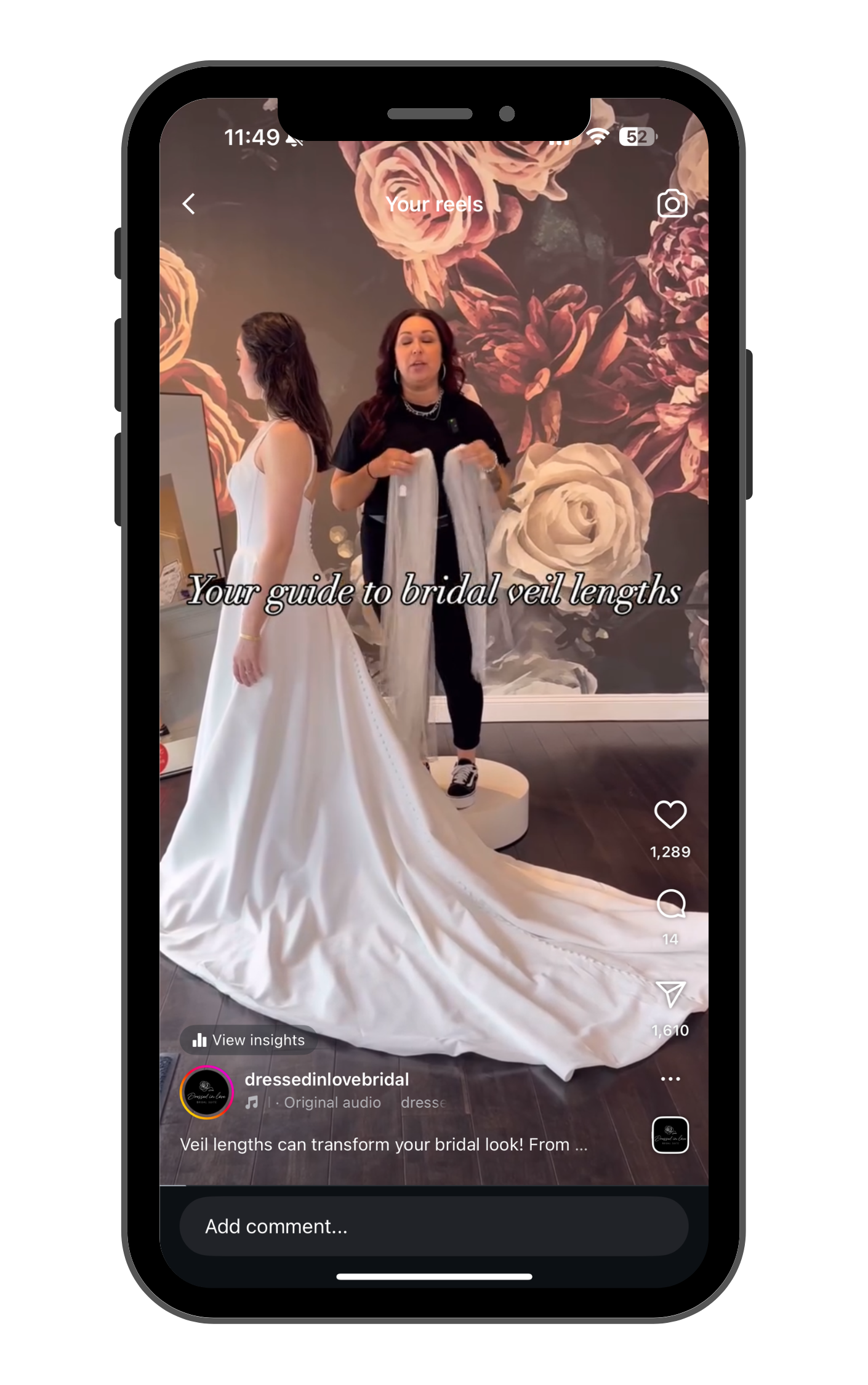 A woman trying on a wedding dress with a long train in a bridal boutique. There is another woman holding veil extensions behind her. The background has dark wallpaper with large pink and white roses.